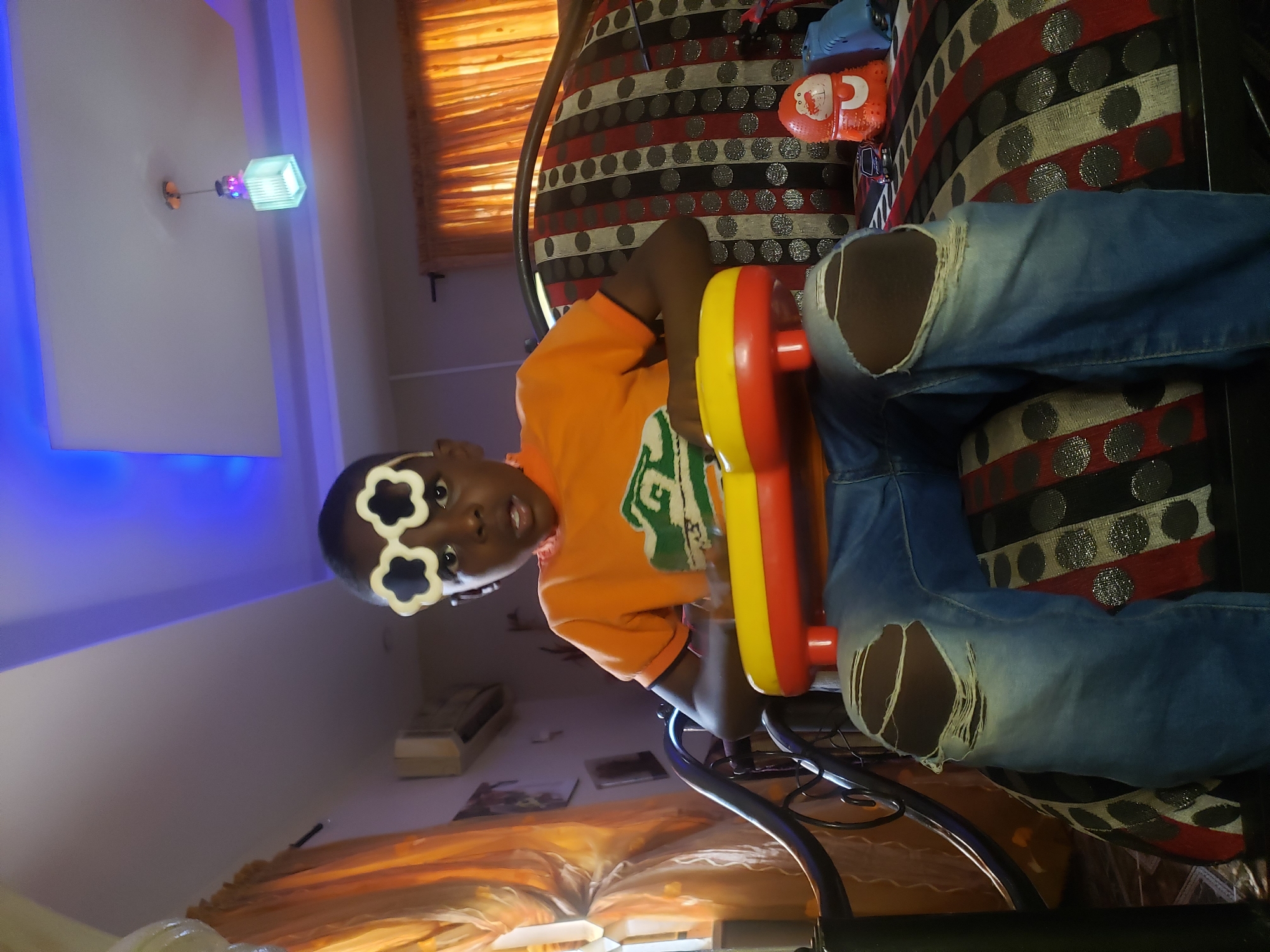 A young Black child wearing torn blue jeans and an orange t-shirt sits on a patterned couch and plays a toy instrument. The child wears white flower-shaped glasses. Behind the child, a window is covered by orange curtains. The room is illuminated by blue lighting in the ceiling and a pendant lamp.