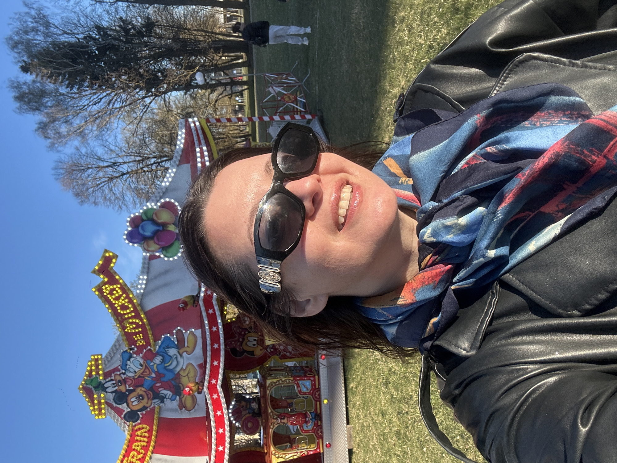 The image captures a vibrant, sunlit outdoor selfie of a woman smiling widely, her face illuminated by bright sunshine. She has dark hair, wears black sunglasses, a colorful scarf with blue, red, and black patterns, and a black leather jacket. Her joyful expression, with a broad smile showing her teeth, suggests a pleasant experience.
Behind her, a colorful funfair attraction is prominently visible. It features a red and white striped design adorned with numerous bright yellow lights. Cartoon characters, specifically Mickey Mouse and Donald Duck, are clearly depicted on the ride's decorative panels. The word "TRAIN" is also visible on the ride's structure. Additionally, colorful balloon-shaped decorations are part of the ride's setup.
The background reveals a large expanse of green grass with several bare-branched trees, suggesting a late winter or early spring season. The sky is a clear, bright blue, indicating sunny weather. A few distant figures of people are scattered across the grassy area, one person notably wearing white pants and a dark top. The overall scene conveys a cheerful, festive atmosphere at an outdoor event, likely a carnival or fair in Riga, Latvia, during the day.