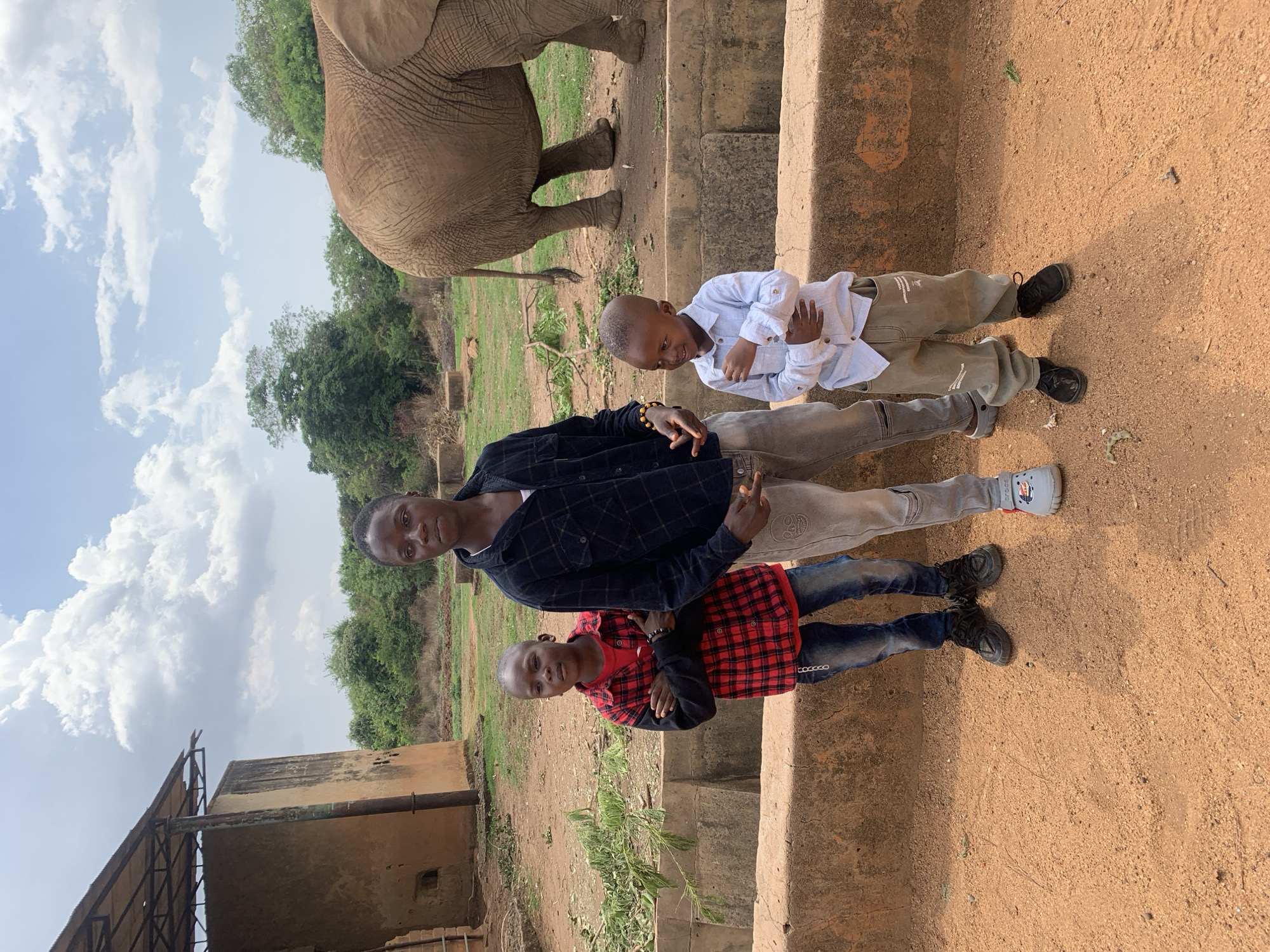 Three boys and part of an elephant standing on a dirt surface.
Location: Jos, Nigeria.
People:
* Three boys of varying ages.
* Youngest: Wearing a red and black plaid shirt, blue jeans and black shoes. Standing with arms crossed.
* Middle: Wearing a black plaid shirt, light colored jeans and blue crocs. Making a peace sign with right hand.
* Oldest: Wearing a light blue shirt, khaki pants and black shoes. Standing with arms crossed.
Subject:
* Elephant: Only the rear of the elephant is visible, including its hind legs, tail and part of its back.
Objects:
* Dirt surface
* Stone barrier with steps
* Building with rusty metal roof
Actions:
* The boys are standing and posing for a photo.
* The elephant is standing behind the barrier.
Interactions:
* The boys appear to be posing for a picture together, possibly with the elephant.
Environment:
* Outdoor setting.
* Vegetation is visible in the background, including trees and grass.
* The sky is visible with some clouds.
* Dirt ground and stone barrier.