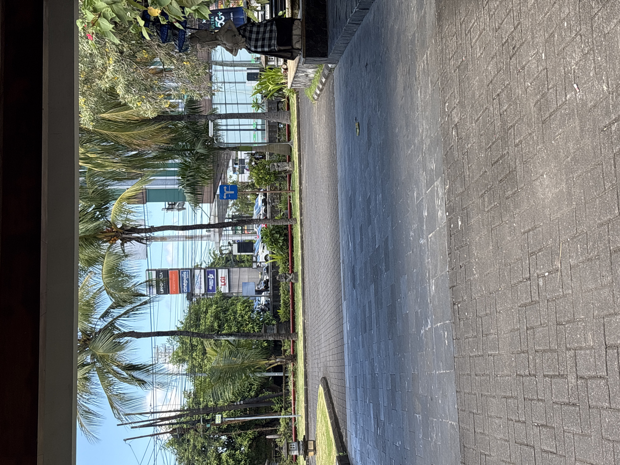 This is an outdoor daytime shot looking out from a partially enclosed space onto a paved area. Beyond the pavement, there is a strip of green grass and tropical vegetation. Palm trees line the street. In the distance, a building with multiple levels and glass windows is visible, along with signs for “Lippo Plaza”, “Hypermart”, “cinepolis”, and “LGI”. Some traffic and pedestrians can be seen in the distance. The general location is South Denpasar, Indonesia.