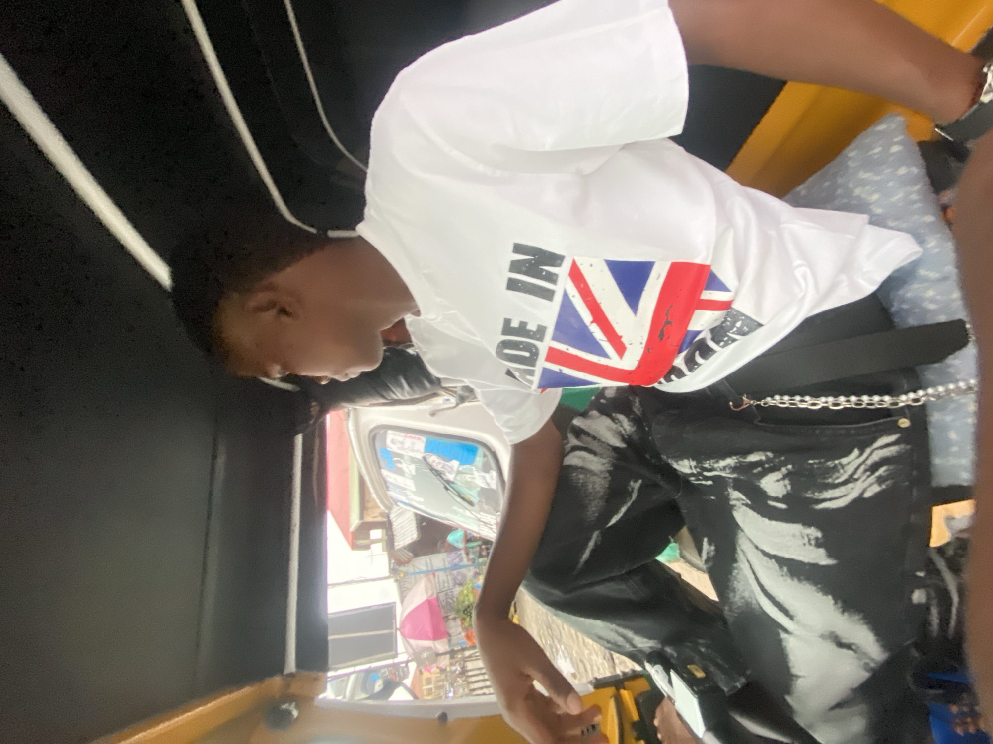 In Ado, Nigeria, a young person sits inside a yellow motorized tricycle. The individual, wearing a white t-shirt with a "Made in the UK" graphic featuring the Union Jack, and black pants with white abstract designs, looks to their right. Their left arm rests on their lap. The interior of the tricycle shows black padding on the ceiling and yellow trim on the sides. Through the open side of the tricycle, a busy street scene is visible, with buildings, a pink and white umbrella, and market stalls. The overall lighting suggests daylight.