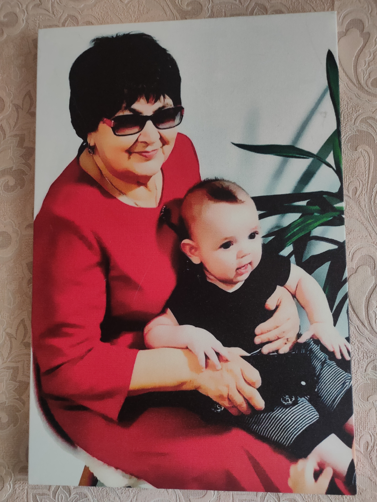 A medium shot portrait depicts a woman and a baby. The woman, with short black hair and dark sunglasses, is wearing a red dress. She is holding a baby who is wearing a black shirt and striped pants. The background consists of a plain light-colored wall and a plant with large green leaves. The image is a printed photograph, possibly on canvas, as indicated by the textured wallpaper visible on the sides of the photograph. The scene is indoors, in what appears to be a domestic setting. The photograph does not provide any visual cues or context related to the city of Oktyabrsky, Russia.