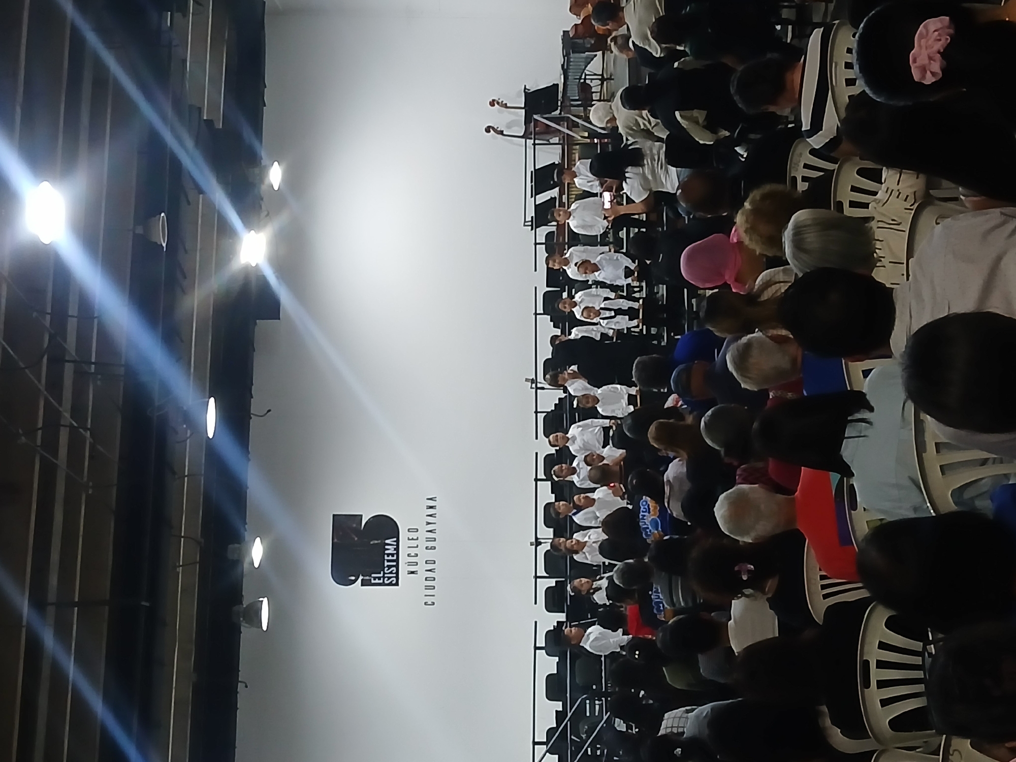 The image depicts an auditorium in Ciudad Guayana, Venezuela, likely a performance space for "El Sistema," as indicated by the large logo on the back wall. The space is filled with people seated in rows of white plastic chairs, facing an orchestra or performance area. The people appear to be an audience watching the performance. The performers are on a raised platform at the far end of the seating, and some individuals seem to be playing stringed instruments. Overhead, lights are affixed to the ceiling beams, and their glare is visible in the photograph. The overall impression is that of a live musical performance taking place in a large public venue.