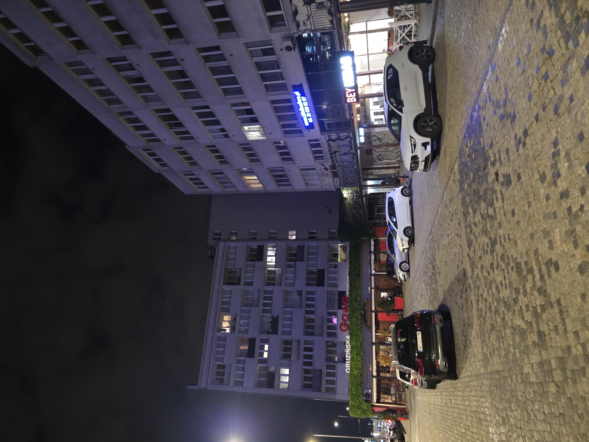 The image captures a street scene at night in Warsaw, Poland. In the foreground, a cobblestone street is visible with two cars parked. The car on the left is a black Mercedes-Benz SUV with license plate WU 2377M. To its right, a white Volvo XC40 SUV is parked.
In the background, there are two multi-story buildings. The building on the left has a facade covered in green ivy above a storefront. A sign above the ivy reads "GRUZIŃSKA GAJA KUCHNIA" with "GAJA" in red lettering. The building on the right is a light-colored apartment building with numerous windows. A blue neon sign on this building displays a website address and a phone number. Below this, a sign reads "BEY KEBAB". The sky is dark with clouds. The scene appears to be a commercial and residential area.