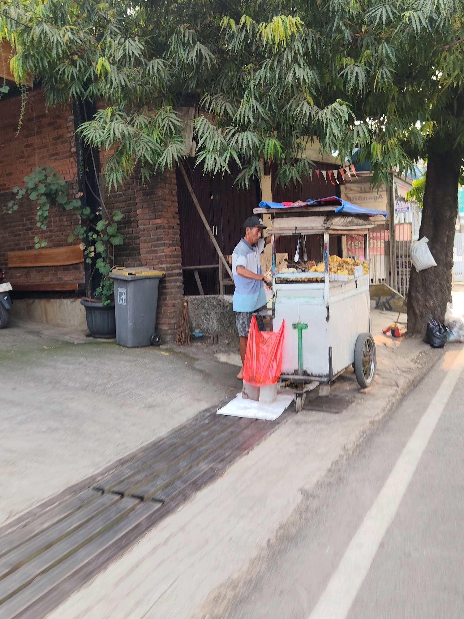 A vendor stands beside a food cart on a sidewalk in Purwakarta, Indonesia. The vendor, wearing a striped t-shirt and shorts, is positioned to the left of the cart, which is laden with food items, possibly fried snacks. A red plastic bag hangs from a stand in front of the cart, and a white tarp covers part of the cart's roof. The background features a brick building and a large tree with dense foliage. A garbage can and a planter are also visible near the building. The sidewalk appears to be made of concrete, and a street with a white line marking runs alongside it. The scene suggests a typical urban setting with street commerce.