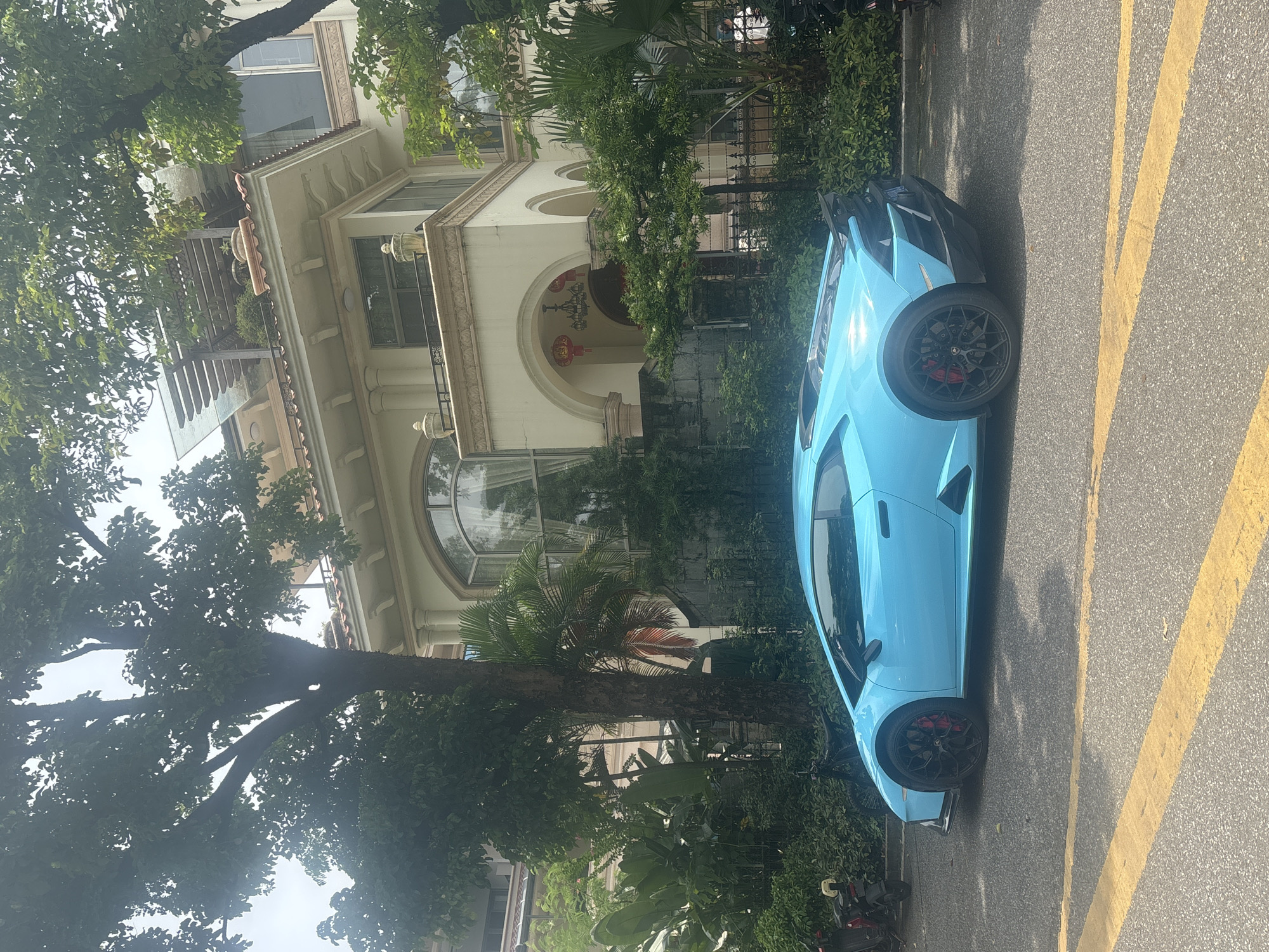 A bright blue Lamborghini is parked on a residential street in Nanhai District, China. The car is positioned on the right side of the frame, facing left, with its rear end closer to the viewer. A large, multi-story building with classical architectural elements, including arched windows and columns, serves as the backdrop. Lush greenery, including trees and bushes, surrounds the building and partially obscures its facade. The street is paved with asphalt and marked with yellow lines. Several motorcycles are visible on the left side of the image, parked near the curb. The sky is overcast, suggesting a cloudy day.