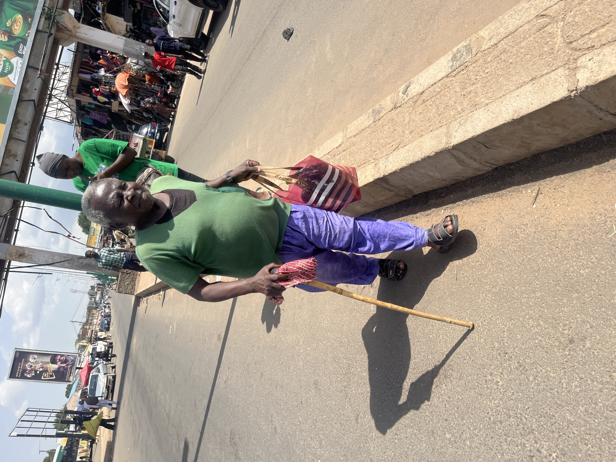 The media file shows a low-angle shot of an older person standing on a paved road, likely in a market area of Jos, Nigeria. The person is wearing a green t-shirt, purple pants, and sandals. They are holding a wooden stick with a red and white patterned cloth wrapped around one end. A translucent red and white striped bag is held in their left hand. The person appears to be looking towards the camera.
In the background, a bustling market scene is visible, with stalls selling various goods, including clothing, and several people are present. Vehicles, including cars and motorcycles, are also seen on the road, suggesting a busy urban environment. A large billboard with text and images is visible on the left side of the frame, as well as another billboard further in the background. The sunlight indicates it is daytime, likely mid-morning or afternoon, and the weather appears to be clear and sunny. The overall mood seems to be one of everyday life in a lively commercial area.