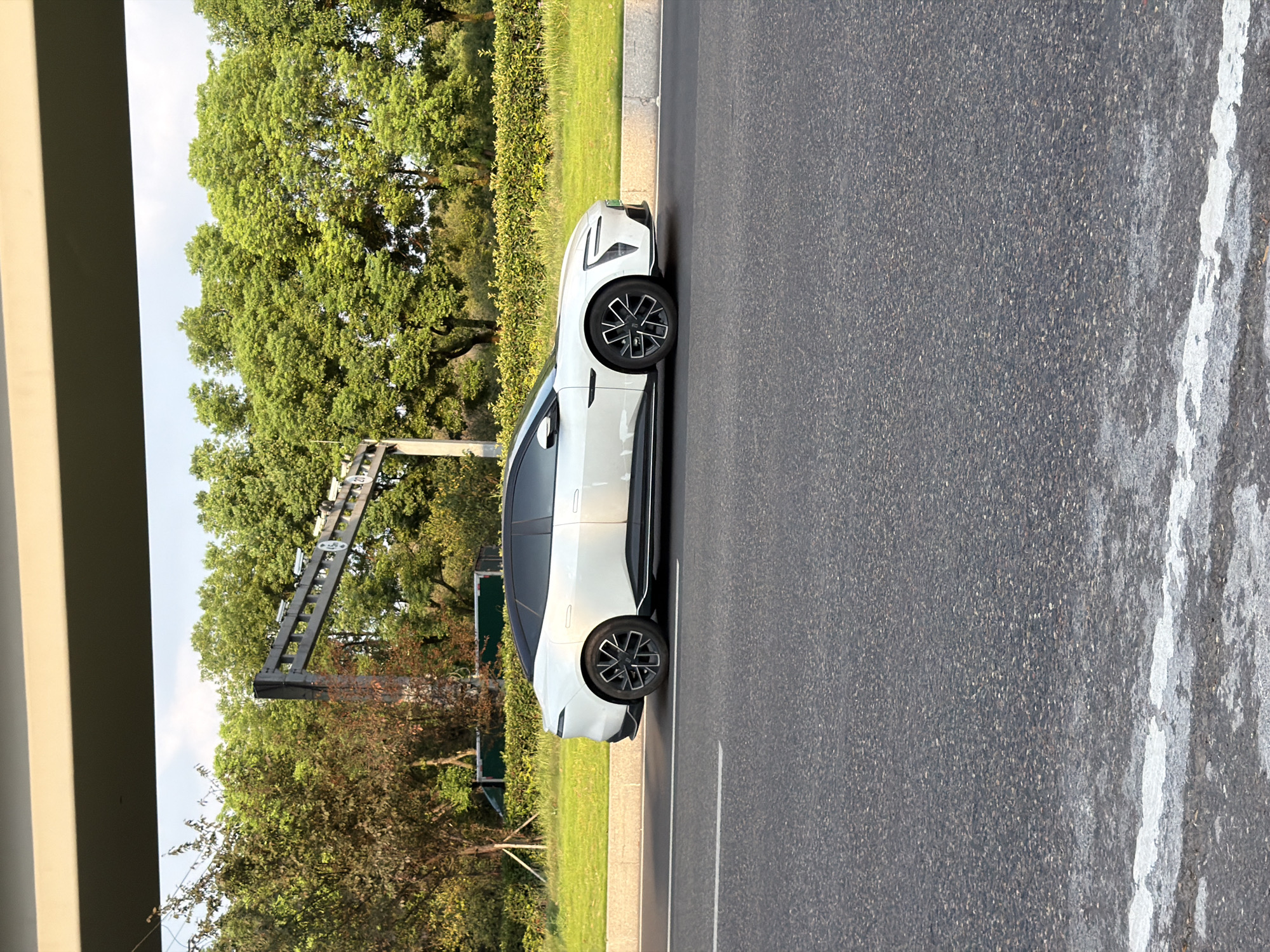 A white electric car is driving on a road in Jiangjin, China. The car is a sedan with a sleek, modern design. It is white with a black roof and tinted windows. The wheels are black and silver with a unique spoke design. The car is in motion, as indicated by the slight blur of the wheels.
The road is paved with asphalt and has a white line marking the lane. To the right of the road is a grassy embankment with a hedge row. In the background, there are trees and a large overhead structure with cameras and signs. The sky is blue with some clouds.
The image appears to have been taken during the daytime, possibly in the late afternoon, as the sunlight casts long shadows. The overall impression is one of a modern city with clean, well-maintained roads and infrastructure.