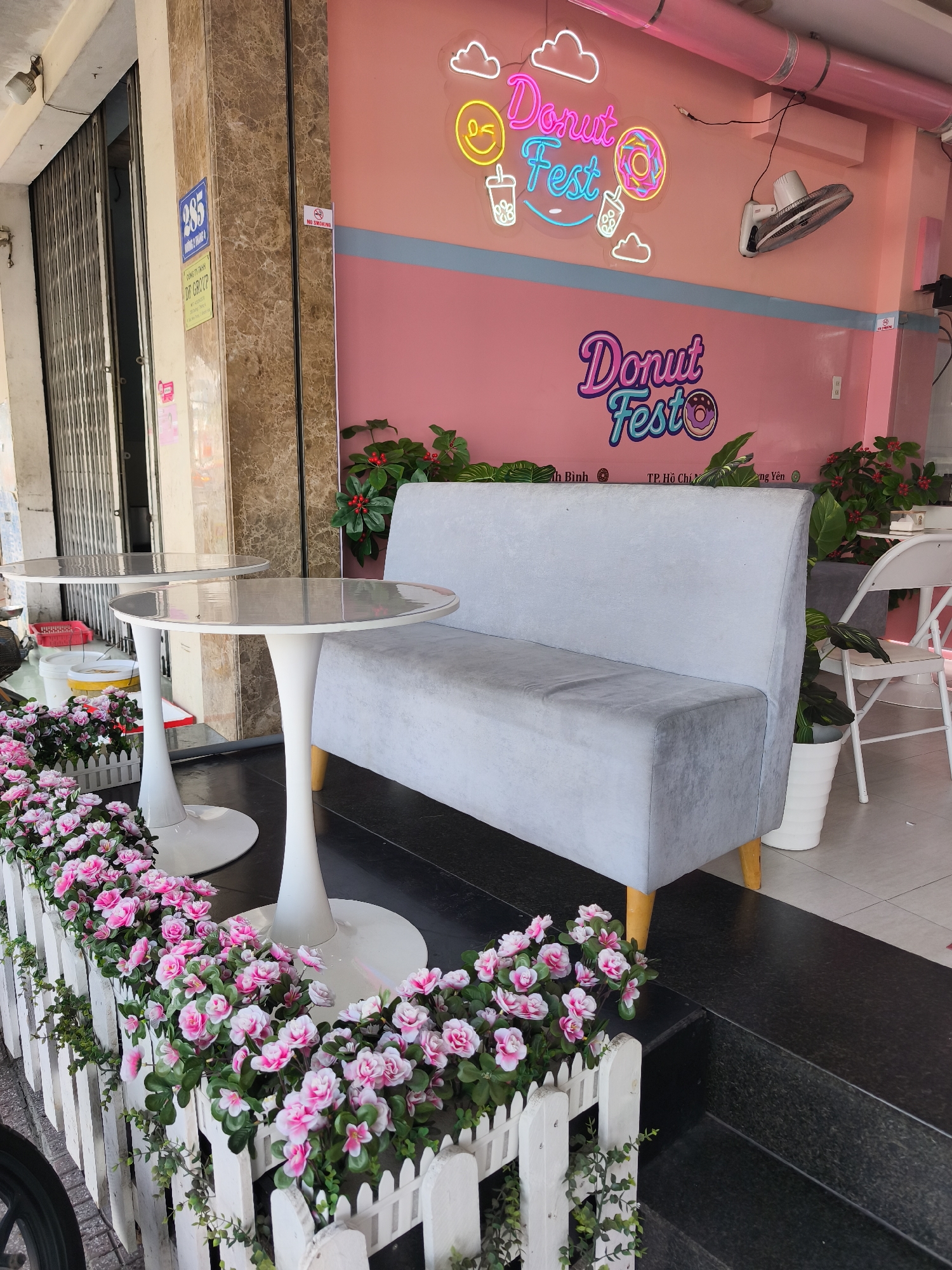 The image depicts the exterior of a cafe, likely located in Bac Nha Trang Ward, Vietnam, as suggested by some textual elements. The facade features a vibrant pink wall adorned with neon signs that spell out "Donut Fest" in a playful, stylized font, accompanied by imagery of clouds and donuts. Below the neon signs, another "Donut Fest" logo is present in a similar font, this time with a donut graphic.
In front of the cafe, two white, tulip-shaped tables with round, reflective tops are positioned. A light gray upholstered bench with wooden legs sits behind the tables. The area is decorated with a low, white picket fence overflowing with an abundance of pink artificial flowers and green foliage. In the background, a closed metal gate and a beige marble-tiled pillar are visible, with a blue sign displaying the number "285" and Vietnamese text. A small red sign with "NO SMOKING" is also visible. A white folding chair and potted plants are seen further inside the cafe area. The ground is paved with dark tiles.