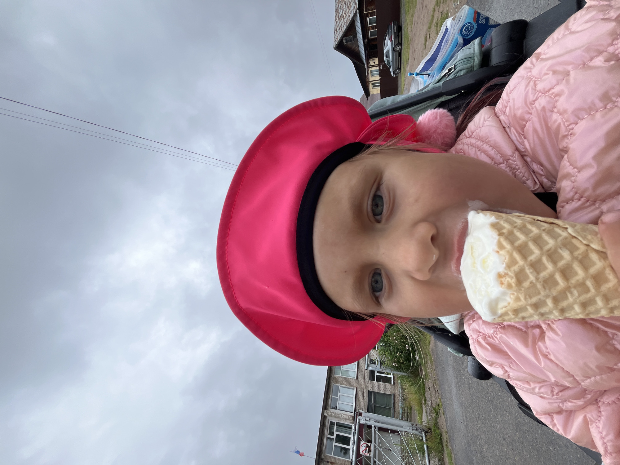 A young girl with blonde hair, wearing a bright pink beret and a pink puffer jacket, is eating an ice cream cone. She is in a stroller, with the edge of the stroller visible in the foreground. In the background, there is a brick building with windows, a metal fence, and a Russian flag on a flagpole. To the right of the building, there is a wooden house with a rusty roof and a car parked in front of it. The sky is overcast. The scene appears to be in a rural or suburban area in Chaikovsky, Russia.