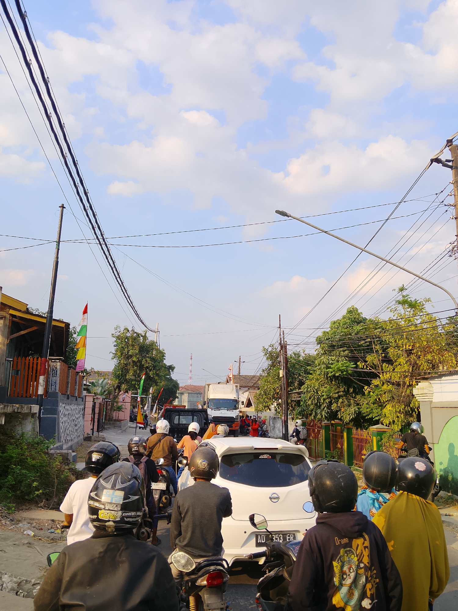 The image depicts a congested street scene in Ngelo, Indonesia, likely during daylight hours given the bright sky and shadows. The sky is a light blue with scattered white clouds. Overhead, numerous black electrical wires are strung between utility poles.
The street is filled with a traffic jam consisting primarily of motorcycles and a few cars. Several individuals are seen riding motorcycles, many wearing helmets, suggesting a common mode of transportation. One white car is prominently featured in the middle of the road. Further down the street, a white truck is visible.
On the left side of the street, there are buildings with wooden fencing and some greenery. On the right, more trees and fences line the road, with a distinct green and white building visible. Several Indonesian flags, in red and white, are displayed on poles, adding a patriotic element to the scene. The overall impression is one of a busy, everyday street in a populated area.