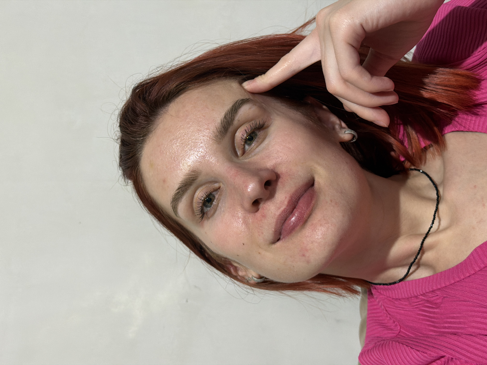 A close-up shot of a woman with reddish-brown hair, light complexion, and light brown eyes. She is wearing a pink top and a thin black beaded necklace. Her right hand is raised, with a finger touching her forehead. She has light pink lipstick on. A small hoop earring is visible on her left ear. The background is a plain light gray/white. No other specific details suggest the location being Chaikovsky, Russia.