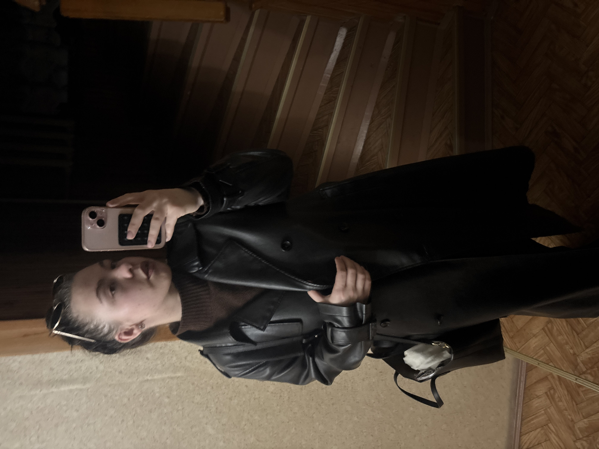 A woman is taking a selfie in front of a staircase. She is wearing a brown turtleneck sweater and a black leather trench coat. She is holding a pink iPhone in her right hand. There is a purse hanging off her shoulder. The staircase has wooden planks and is illuminated with dim lighting. The walls appear to be paneled with wooden slats. The floor has a herringbone pattern. The location is Chaikovsky, Russia.