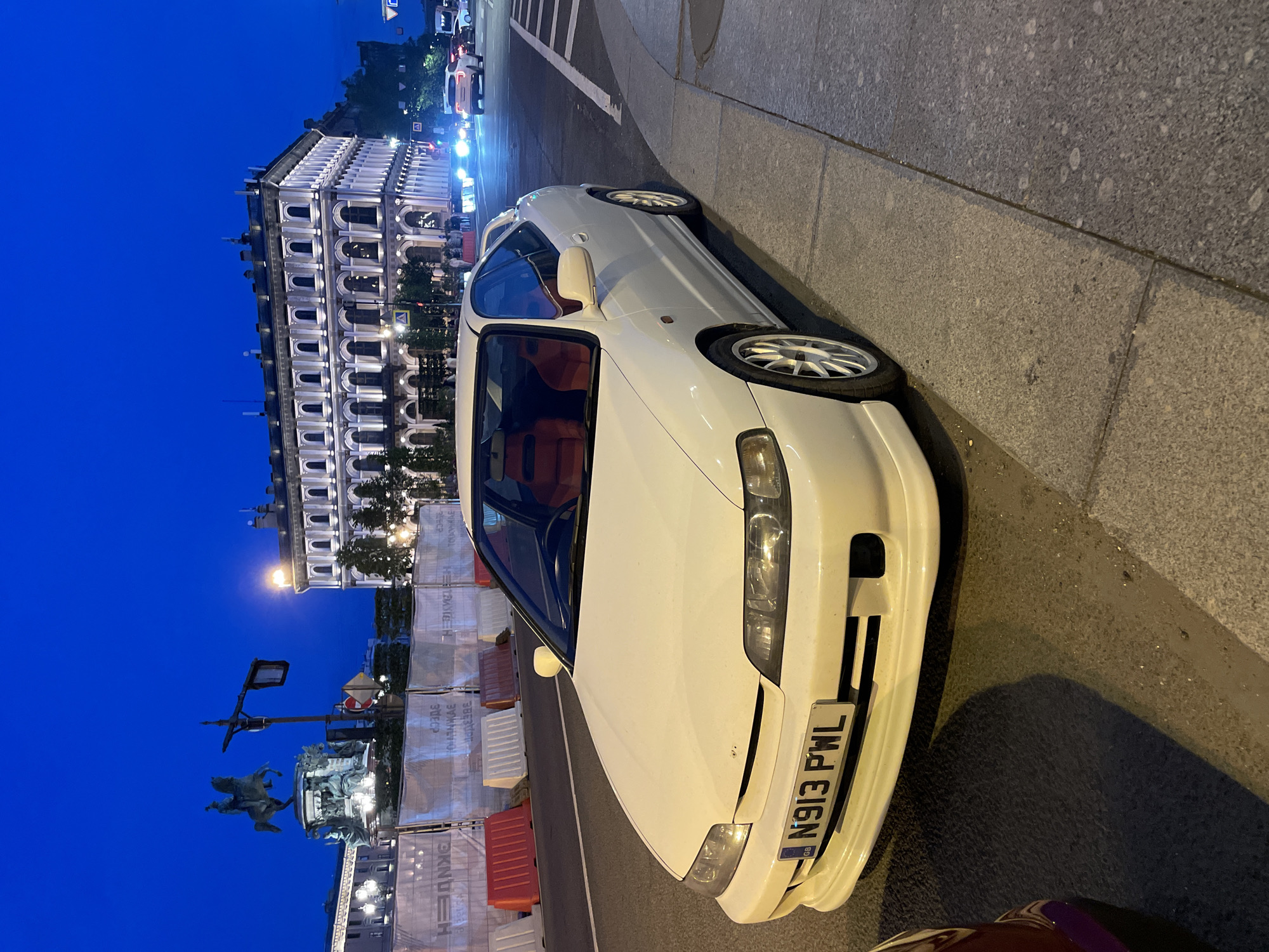The image depicts a white Honda Integra DC2 coupe parked on a street at dusk in Vaganovo, Russia. The car is positioned in the foreground, facing slightly to the right, with its front bumper and driver's side visible. The vehicle has a British license plate that reads "N913 PWL" and red interior seats.
Behind the car, a row of white and red construction barriers lines the sidewalk. In the middle ground, a large, ornate building with multiple arched windows is illuminated, suggesting it is a historical structure or a place of importance within the city. A street light and a traffic signal are visible near the building. Further in the background, an equestrian statue on a pedestal stands against the twilight sky. Other vehicles, including a white SUV and a dark car, are visible in the distance on the road, indicating traffic activity. The sky is a deep blue, transitioning from the day to night. The overall environment suggests an urban setting, likely a public square or a busy street in Vaganovo. There are no discernible people in the immediate vicinity of the car, though some figures may be present near the illuminated building.