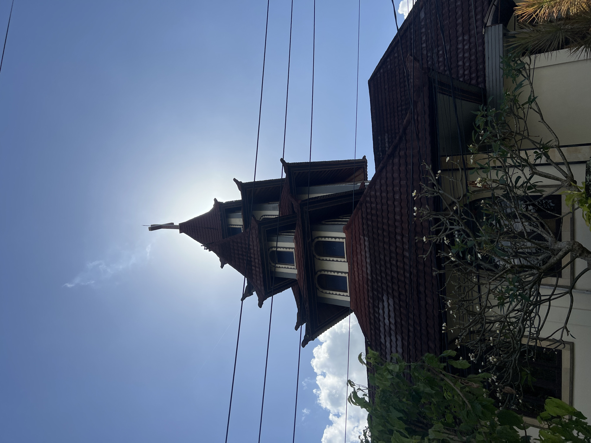 The image shows the roof and upper levels of a building, likely a church or a temple, with a distinct tiered roof structure and multiple gables. The building is seen from a low angle, looking up towards the sky. The sun is bright and positioned behind the upper part of the structure, creating a lens flare effect. The sky is a clear blue with some scattered white clouds. Several dark electrical wires run horizontally across the frame in front of the building. In the foreground, there are branches of trees with green leaves and a tree with small white flowers. The building itself has a dark tiled roof and visible architectural details such as arched windows and decorative eaves. The overall scene suggests an outdoor setting on a sunny day.