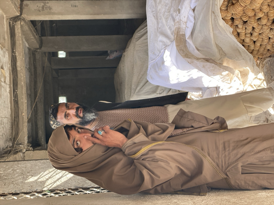 The image displays two males inside a concrete building, consistent with a market or storage area, located in Kandahar, Afghanistan. In the foreground, positioned to the left, a young male with dark hair and a light complexion wears a brown headscarf and a matching long-sleeved garment adorned with gold trim. He has a prominent turquoise ring on his right ring finger and is looking directly at the camera, with his right index finger touching his nose. Behind him and slightly to the right stands an older male with a grey beard, wearing a patterned skullcap, a light-colored patterned shirt, and a dark vest. He is also looking directly at the camera. The background features concrete pillars, a concrete ceiling with visible electrical wiring, and large piles of items covered with white and beige tarps. In the lower right, a stack of circular, coiled, light-brown, woven materials is partially visible. Natural light illuminates the scene from the left.