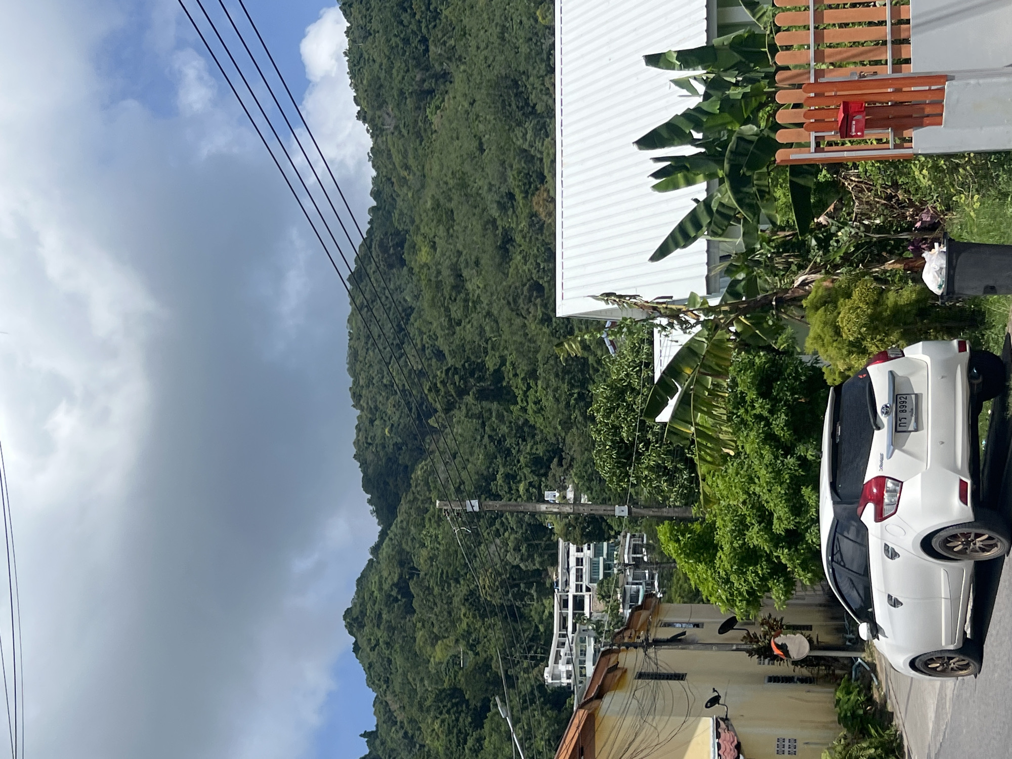 In Choeng Thale, Thailand, a white hatchback car is parked on the side of a road. The car faces left and has a license plate with Thai characters and the number "กจ 8992". The car is in front of a residential area with a pale yellow building on the left, featuring an orange-tiled roof and satellite dishes. Behind the yellow building, modern white multi-story structures are visible, nestled into a lush green, tree-covered hillside. A white corrugated metal roof of another building is prominent in the foreground to the right, with banana trees and other foliage. A wooden fence with vertical slats, painted orange, encloses the property on the right, and a red mailbox is attached to it. A black trash can with white plastic bags is situated near the fence. Overhead, power lines stretch across the frame against a sky with a mix of gray clouds and patches of blue. The overall scene depicts a suburban or semi-rural residential area within a tropical climate.