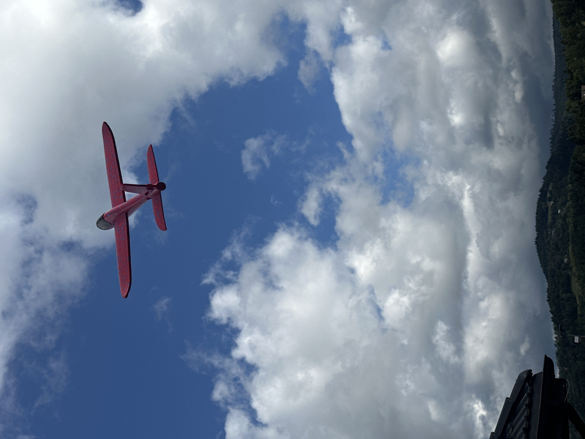 An overhead shot captures a bright red model airplane soaring against a vibrant blue sky dotted with fluffy white clouds. The airplane, with its wings extended, is positioned centrally in the frame, angled slightly downwards. In the lower part of the image, a dark roofline is visible, with a green, tree-covered hillside stretching into the background. The overall scene suggests an outdoor activity, possibly in a park or open area, with the landscape indicating a location with natural topography. The city of Wisła, Poland, is known for its mountainous terrain and recreational areas, making this image consistent with such a setting.