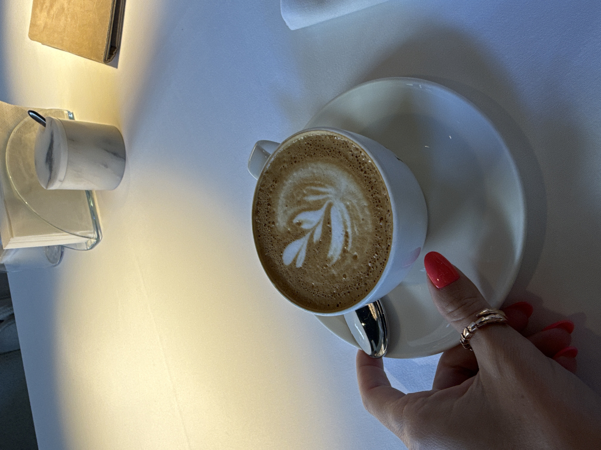 A hand with bright red nail polish and a gold ring is holding a spoon next to a white cup of coffee with latte art. The cup sits on a white saucer on a white tablecloth. In the background, there is a marble sugar dispenser with a small spoon and a folded menu or book. The scene is set on a table that appears to be in a restaurant or cafe. There is no indication of the location being in Moscow, Russia, nor any specific details that would suggest it.