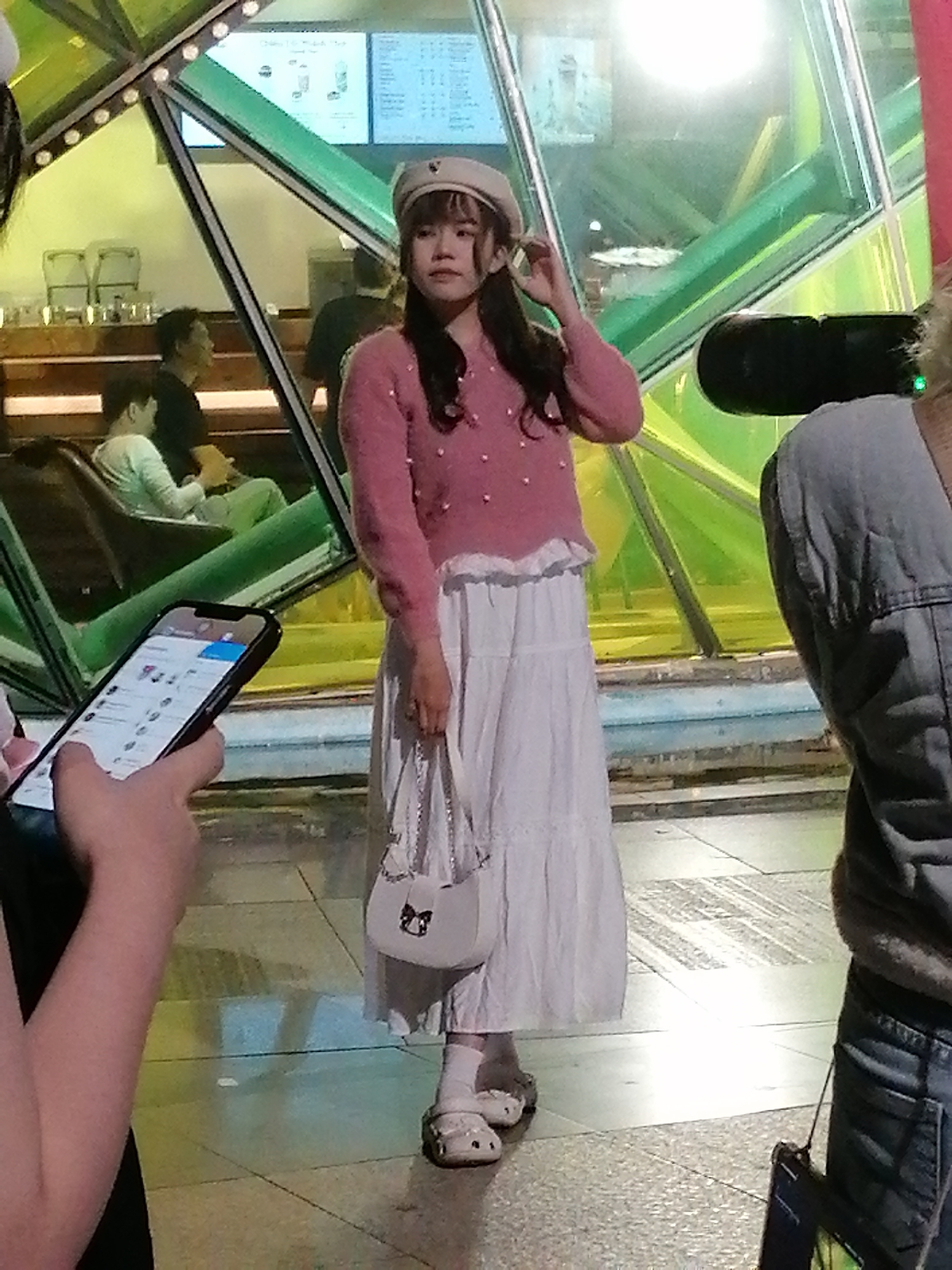 A young East Asian woman wearing a pink knitted sweater, a white tiered skirt, and a white beret stands in front of a brightly lit, modern architectural structure with green and yellow tinted glass. She is holding a small white purse with a chain strap and a cartoonish black bow on the front. Her right hand is touching her beret. She is also wearing white socks and white clogs with a cartoonish face on them. In the background, there are blurred figures of people sitting on chairs and looking at their phones. A hand holding a smartphone is visible in the lower left of the frame, with the screen displaying a social media feed. The scene appears to be indoors, possibly in a cafe or a public waiting area, with tiled flooring and bright artificial lighting. The location is identified as Da Lat, Vietnam.