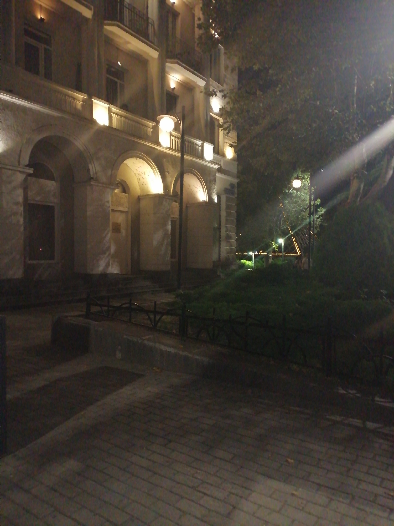 An exterior nighttime street-level photograph shows a building with arched entrances and balconies. The building's facade is illuminated by artificial lights, highlighting architectural details. To the right of the building, a streetlamp illuminates a grassy area with trees and a pathway. The foreground is a brick-paved sidewalk. The overall scene depicts a quiet urban environment at night.