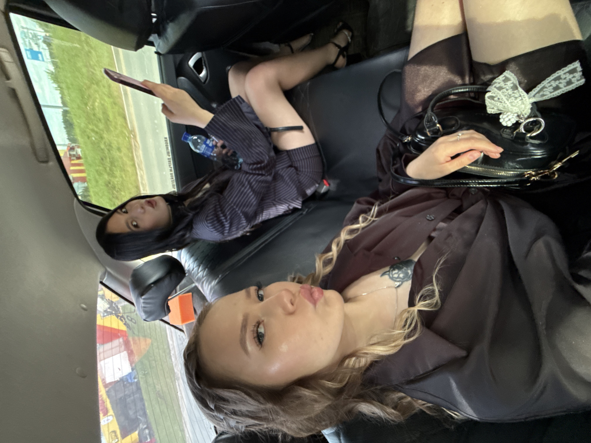 Two young women are seated in the back of a vehicle, likely a taxi or personal car. The woman in the foreground, with blonde curly hair styled in a braid, is looking directly at the camera with her lips pursed. She is wearing a dark, possibly satin, blouse and a necklace with a pendant. A black handbag with a white ribbon charm is visible in her lap. The woman behind her, with long dark hair, is looking at her phone, which she is holding in her right hand. She is wearing a pinstripe suit with a short skirt and high heels. A bottle of water is in her left hand. The interior of the car appears to be standard, with black leather seats and a headrest. The windows show a view of a roadside with green grass and trees, suggesting a location outside of a densely populated urban area, possibly on the outskirts of the city of Chaikovsky, Russia, given the provided context. The overall impression is a casual snapshot taken during a car ride.