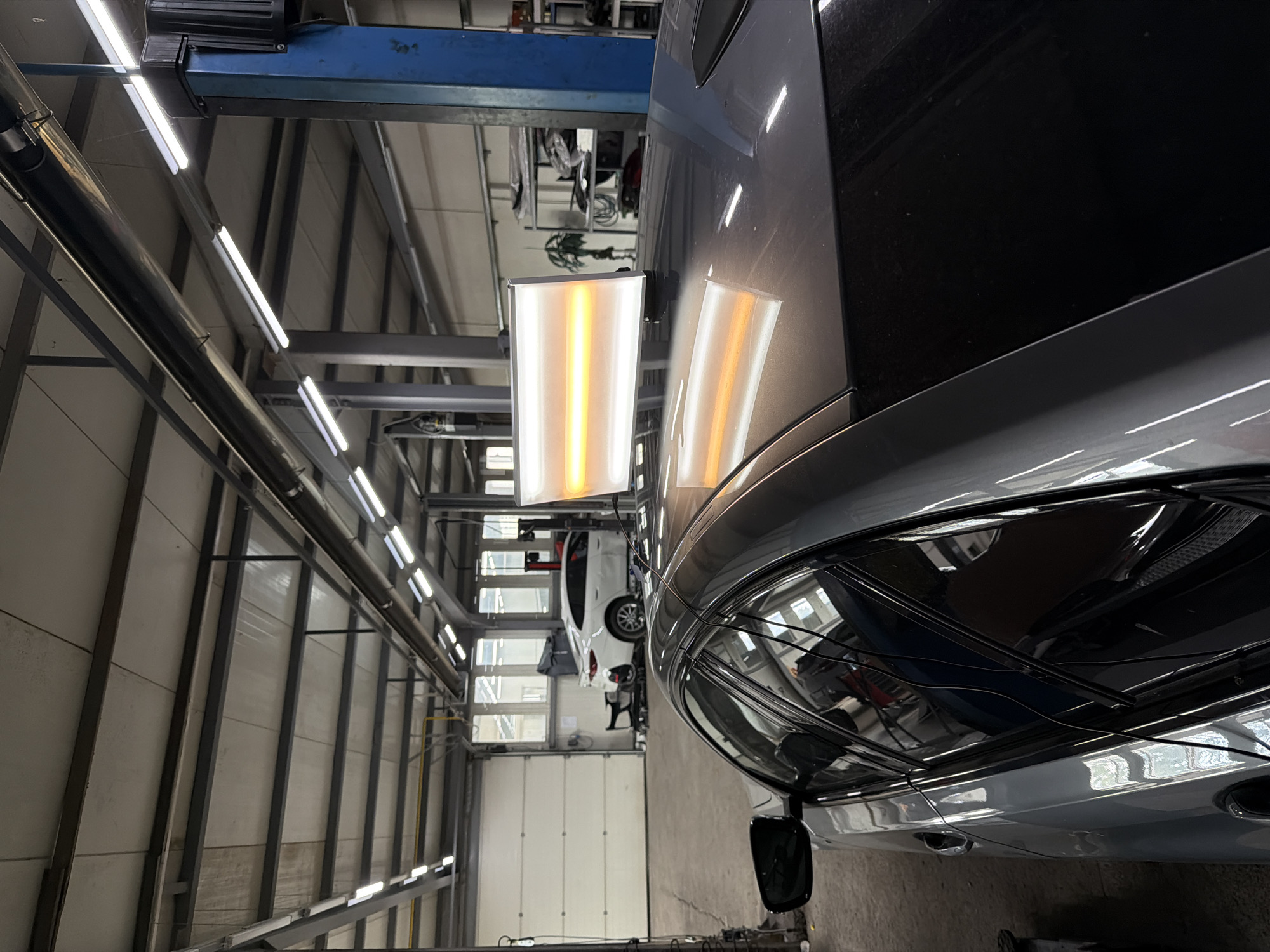 This is a close-up, eye-level, interior shot of a car repair shop in Burgas, Bulgaria. The foreground features the roof and passenger side of a dark gray sedan, with a PDR (Paintless Dent Repair) light bar mounted on the roof, casting orange and white reflections. The background shows a white garage door, a white sedan parked behind it, shelving units with items, and a blue car lift. The ceiling is industrial, with exposed beams and fluorescent lighting. The floor is concrete. There are no people visible in the image.