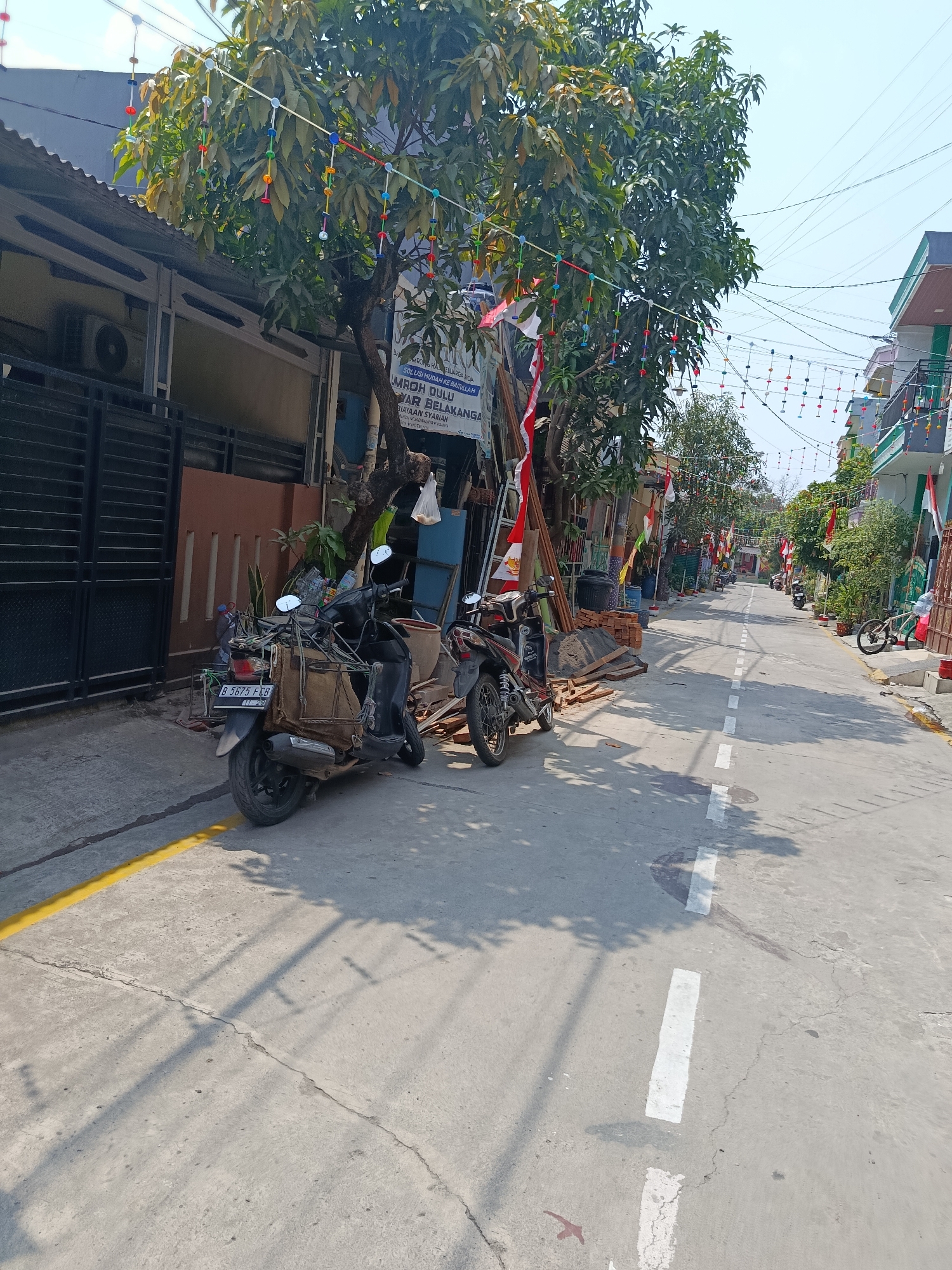 The image depicts a street scene in Wanasari, Indonesia, likely during a festive or celebratory period, indicated by numerous Indonesian flags and colorful decorative garlands strung across the street and from trees. The street is paved, with white dashed lines marking the center. On the left side of the street, a dark-colored metal gate is visible, adjacent to a building with an air conditioning unit mounted high on its exterior. A motorcycle, loaded with bags and other items, is parked on the left side of the street, with its license plate reading "B 5675 FCB". Further down the street, several other motorcycles and a bicycle are visible. Buildings line both sides of the street, some with balconies. Trees with dense green foliage are interspersed along the street, and construction materials, including bricks and lumber, are stacked on the side of the road. The sky is clear and bright, suggesting it is daytime. Shadows of trees and power lines are cast on the street, indicating the sun's position. The overall impression is of a residential or commercial street decorated for a local event.