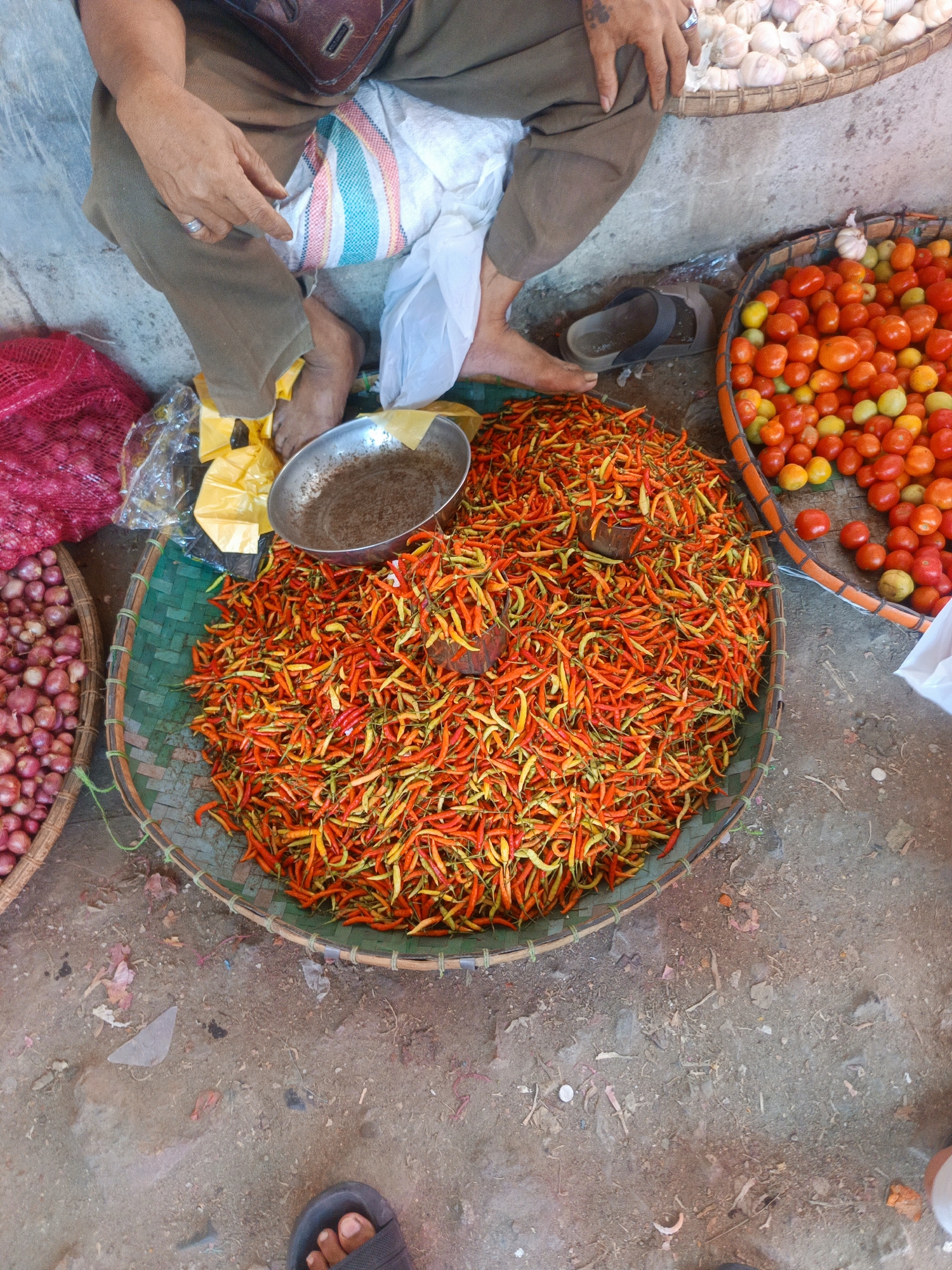 This is a high-angle, close-up shot of a market stall in Gorontalo, Indonesia. A person, wearing brown pants and a ring on their left hand, is seated. In front of them is a large, round, woven basket overflowing with small, brightly colored chili peppers, ranging from yellow to orange to red. To the right of the chili peppers is a round basket filled with ripe tomatoes, various shades of red and orange, with some green tomatoes mixed in. To the left of the chili peppers, partially visible, are red onions in a woven basket and a pink mesh bag also containing red onions. A small metal bowl containing a dark powder is placed on top of the chili peppers. The ground is dirt and scattered with debris. Another person's bare foot in a dark sandal is visible in the bottom of the frame.
