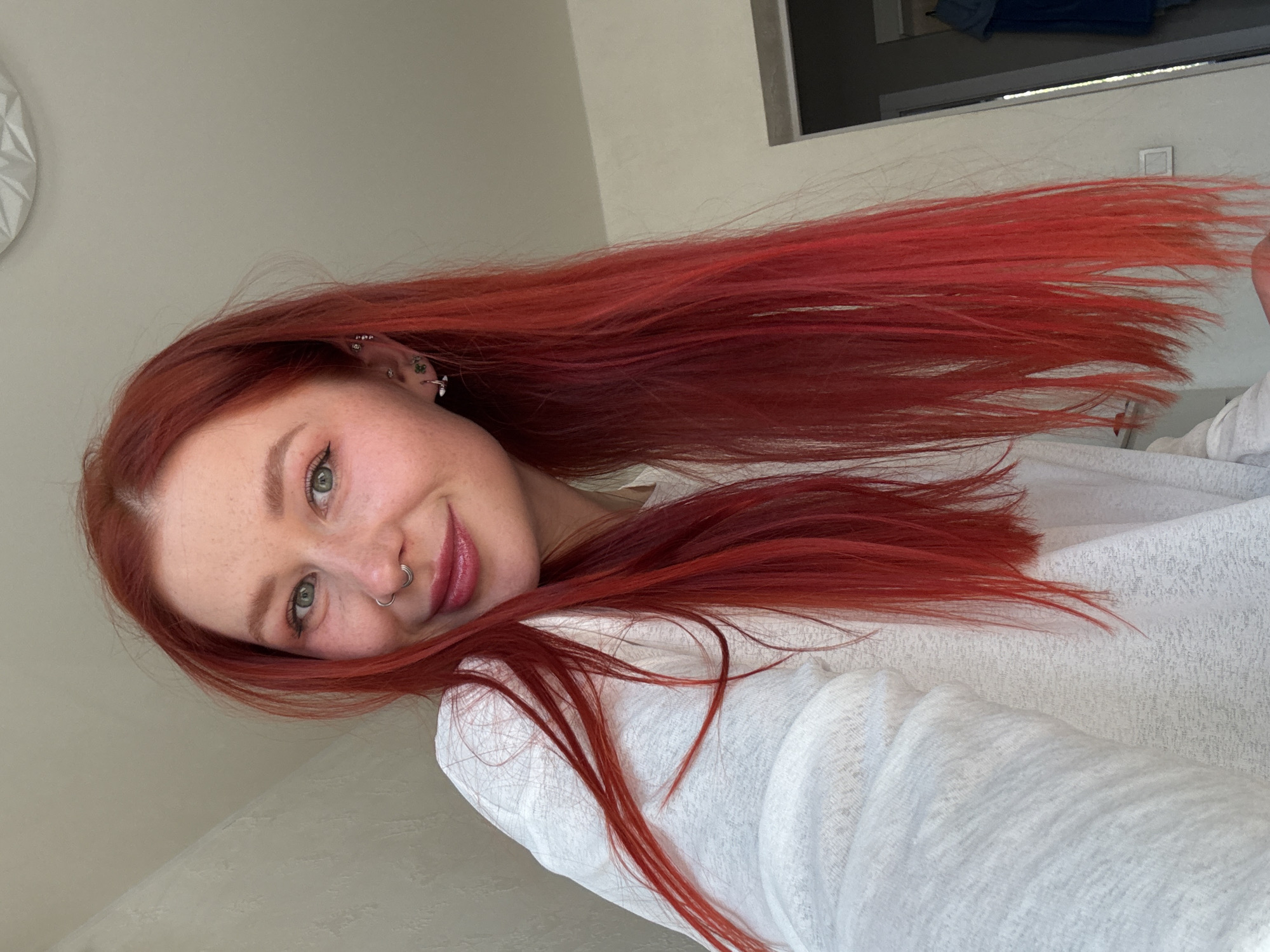 A young woman with striking, long, vibrant red hair is the central subject of this close-up portrait, oriented diagonally across the frame. Her head is positioned in the lower-left, and her abundant hair cascades across the upper and right portions of the image. She has fair skin with visible freckles scattered across her nose and cheeks. Her eyes are a distinctive light green, and she is looking directly at the camera with a gentle, closed-mouth smile, conveying a pleasant and content expression.
Notable facial features include multiple silver piercings on her visible left ear, including helix, tragus, conch, and lobe piercings. She also has a silver septum ring piercing her nose. She is wearing a light-colored, possibly off-white or very light grey, long-sleeved top made from a textured, somewhat sheer fabric.
The background suggests an indoor setting, likely a room in a home in Rostov-on-Don, Russia. A light-colored, plain wall takes up most of the background. On the far left, a decorative white object with geometric patterns is partially visible. In the upper right corner, a dark-framed window or doorway shows a glimpse of a dark blue or grey object, possibly an item of clothing or a bag, beyond it. The lighting is bright and natural, indicating it is likely daytime. There is no visible text in the image.