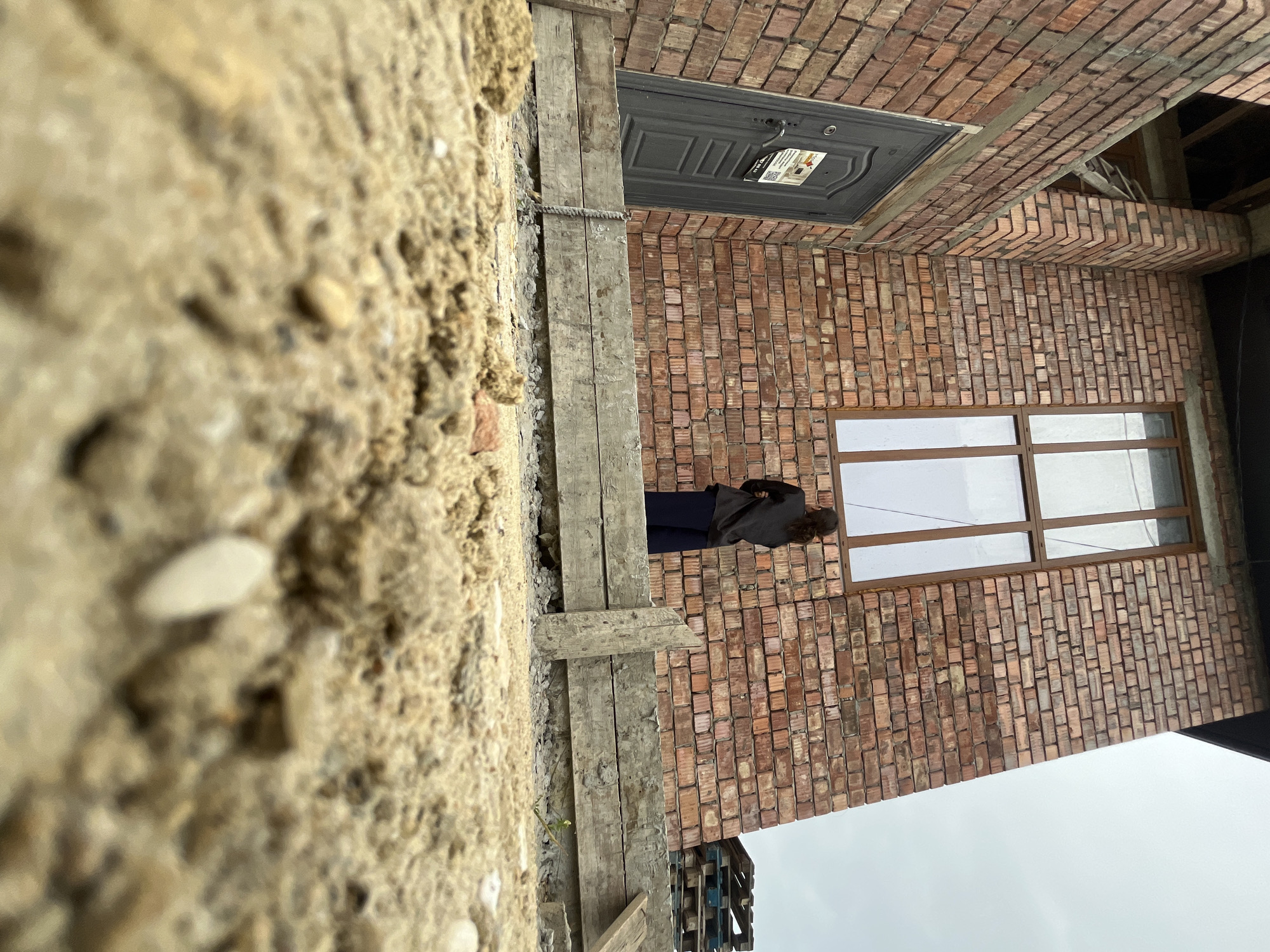 A low-angle photograph captures a brick building, likely under construction or newly completed, in Kaspiysk, Russia. The immediate foreground is a blurred expanse of sandy soil interspersed with small pebbles. A rough wooden barrier, possibly formwork or a temporary step, spans the midground. Behind this barrier, a person viewed from the back stands on a gravelly surface, facing the building's red-brown brick facade. The individual wears a dark top and dark trousers, positioned directly beneath a large, multi-paned window with brown frames. To the left, a dark gray metal door with a visible paper notice is recessed into the brickwork. Exposed wooden structural beams are visible above the door, indicating unfinished elements of the building. Stacked wooden pallets are present on the ground to the right of the building, and the sky above is uniformly overcast and light gray.