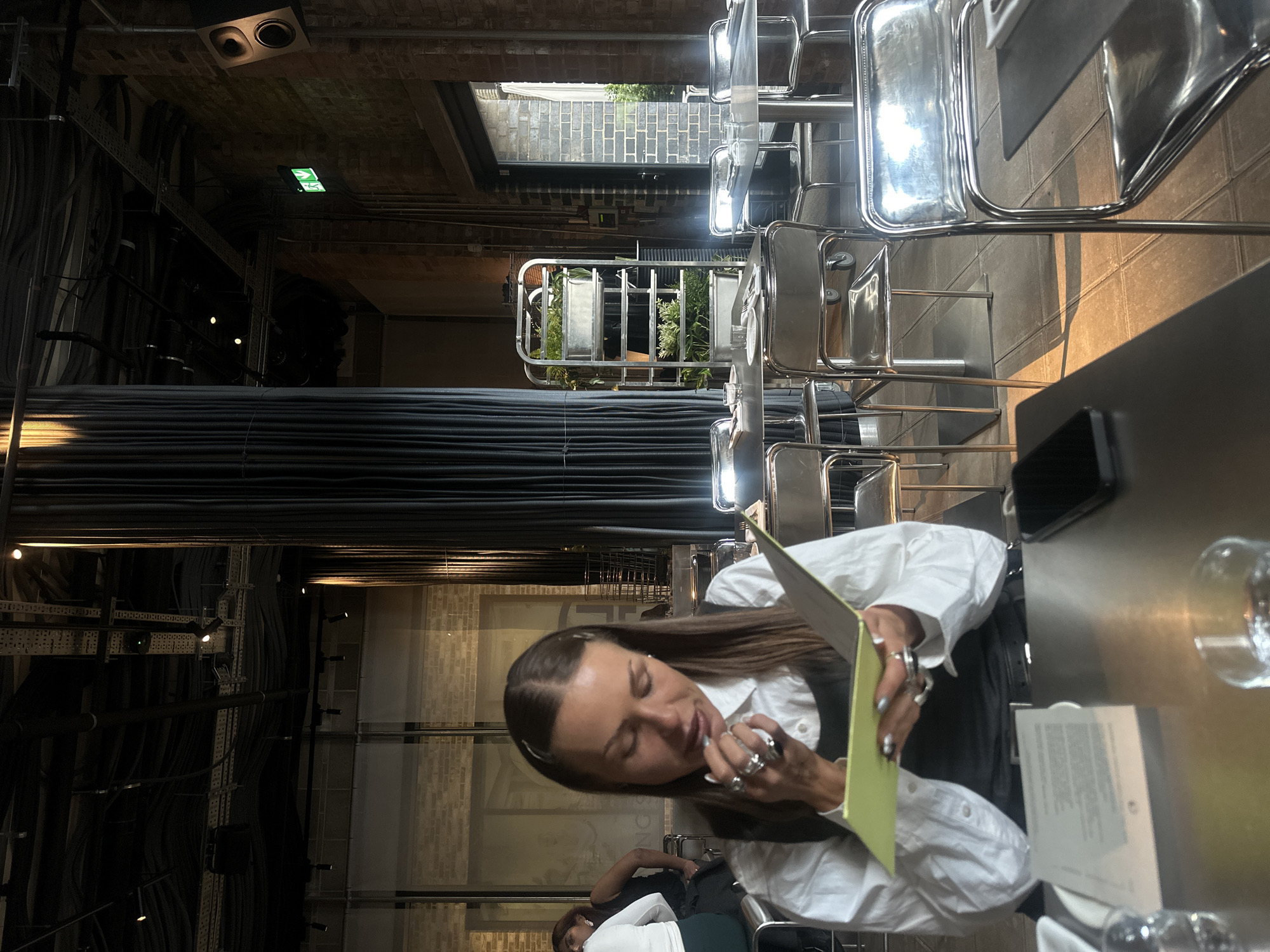 The image shows the interior of a restaurant or dining establishment, likely in London, United Kingdom. A woman with long brown hair, wearing a white collared shirt over a dark vest, is seated at a metal table, looking at a yellow menu. She has multiple rings on her fingers and her nails are painted. A black mobile phone and a glass are on the table beside her. Other people are seated in the background. The interior of the location shows exposed pipes and brickwork, suggesting a modern industrial design. Sunlight filters through an open-air ceiling element or window. Silver chairs are arranged around the tables.