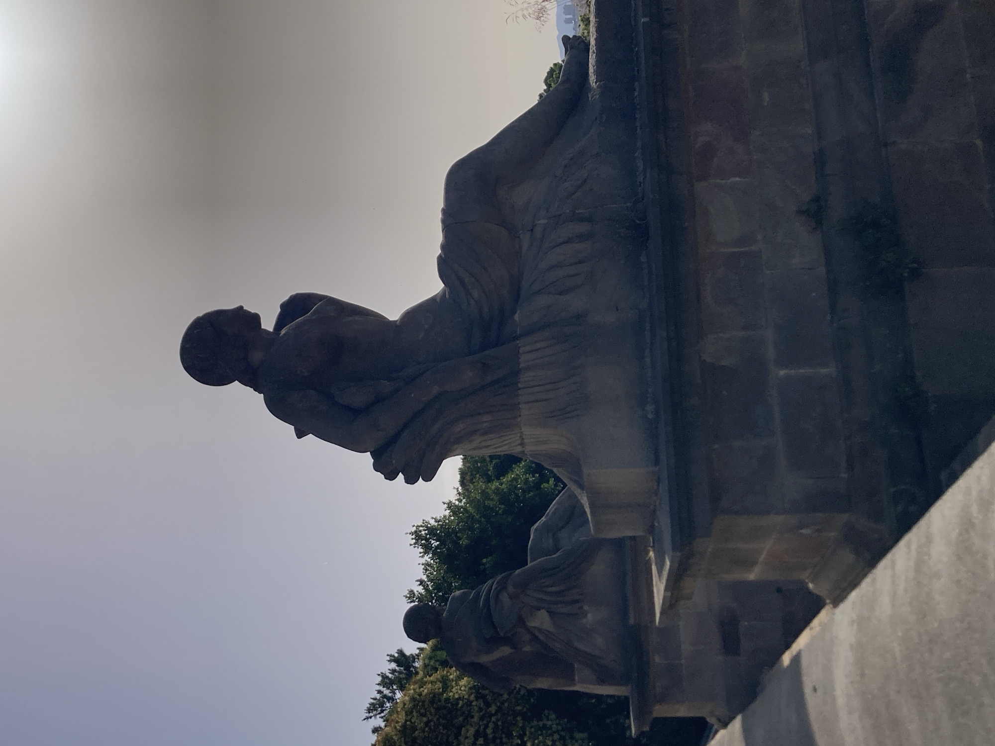 The image shows two stone sculptures of reclining figures, silhouetted against a bright sky, located in Barcelona, Spain. The sculpture in the foreground is more detailed, depicting a female figure with draped clothing and her body turned to the right. The second sculpture, further back and to the left, appears to be a similar reclining figure. Both sculptures are situated on stone pedestals with visible brickwork. Lush green trees are visible behind the sculptures, and in the far distance, faint structures of a cityscape can be seen, including what appears to be a tall building with antenna-like elements. The overall scene suggests an outdoor setting, possibly a park or a monument. The lighting is strong, creating a silhouette effect on the sculptures.