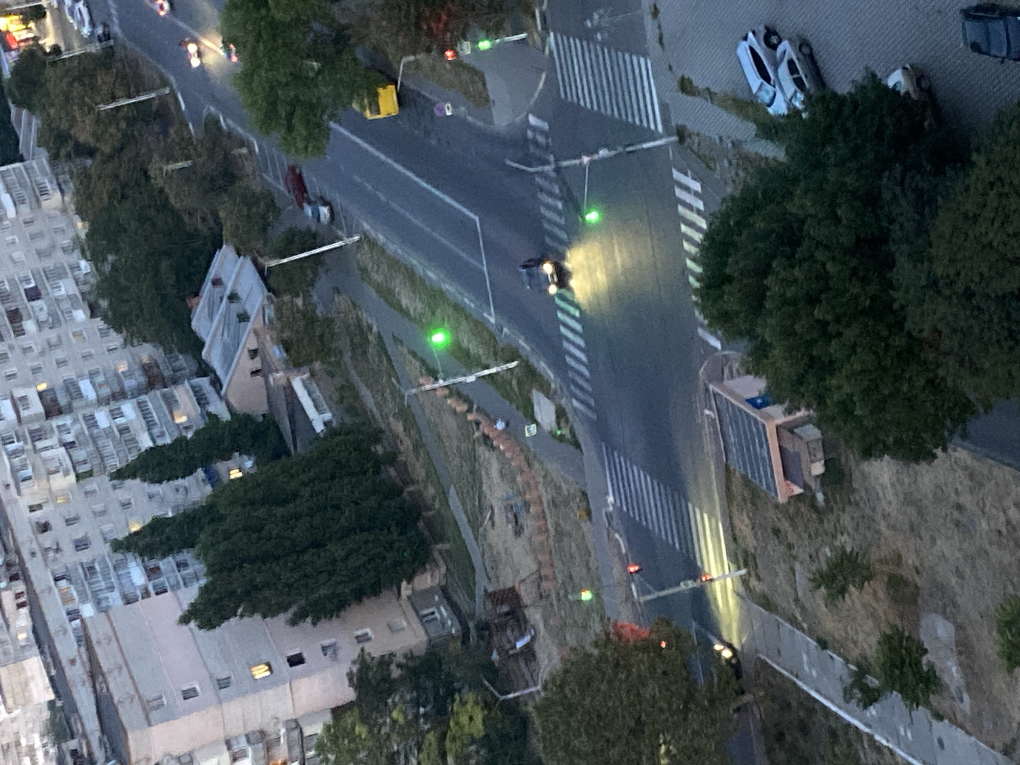 This is an aerial, high-angle shot of an urban street intersection in Chornomorsk, Ukraine, at dusk or dawn. The scene is dominated by residential buildings with balconies and parked cars, interspersed with mature trees. A road with marked lanes and pedestrian crossings cuts through the frame, with traffic lights visible at the intersection. A dark-colored car with headlights illuminated is in the process of crossing the intersection. A yellow bus is parked on the right side of the road. There are no clearly identifiable people in the immediate vicinity of the road. The overall impression is of a quiet urban residential area in twilight.