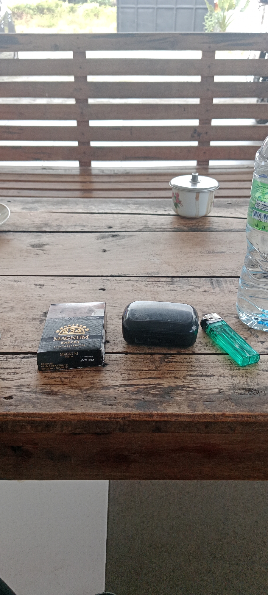 The image displays a close-up view of a wooden table with several items on it, suggesting a relaxed setting, possibly in Tuban, Indonesia. In the foreground, there is a pack of "MAGNUM KRETEK" cigarettes, a black earbud case, and a bright green lighter. Behind these items, a small white ceramic bowl with a lid, decorated with floral patterns, is visible. To the right, a partially visible plastic water bottle with a label indicating it is from Indonesia. The background shows a slatted wooden structure and some greenery, consistent with an outdoor or semi-outdoor environment in Tuban. The lighting suggests it is daytime. There are no people or animals present in the image.