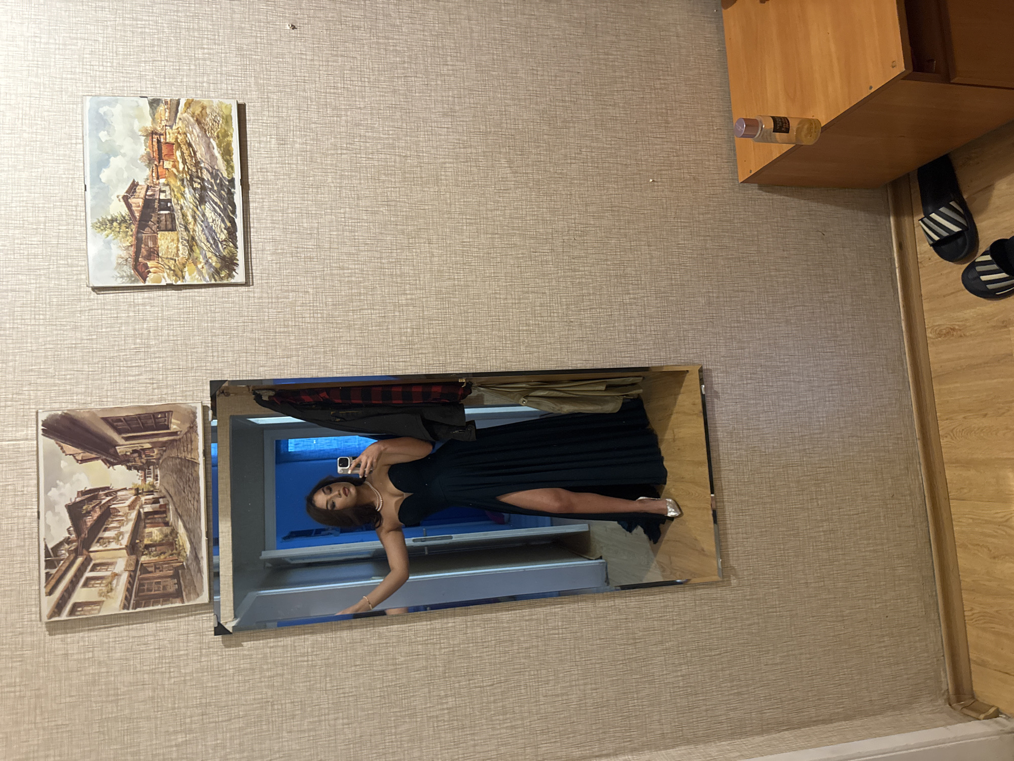 A vertical medium shot shows a room with beige-colored textured wallpaper. On the left, two framed landscape paintings hang stacked. To the right, a wooden cabinet is visible in the upper corner, with a bottle resting on its edge, and two pairs of black-and-white striped sandals below. The main subject is a large rectangular mirror reflecting a woman wearing a long black dress with a high slit, silver heels, and a pearl necklace. She is holding a phone in front of her face, presumably taking a picture in the mirror. A blue-toned closet interior is visible in the background behind her. The location is Unknown, Unknown.