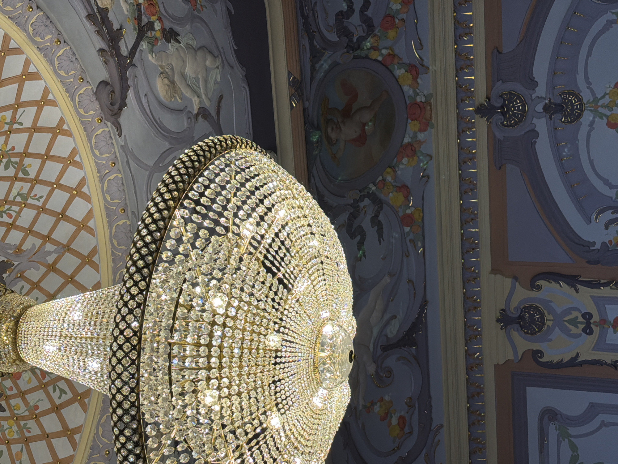 The image shows a close-up of a large, ornate crystal chandelier hanging from a highly decorated ceiling. The chandelier is made of many faceted crystals that reflect light. The ceiling features intricate plasterwork with floral motifs, cherubs, and geometric patterns. The colors are predominantly pastel blues, purples, yellows, and grays, with accents of gold. The overall impression is one of opulence and grandeur. This scene is located in the Nizhny Novgorod State Academic Opera and Ballet Theatre, Russia.
