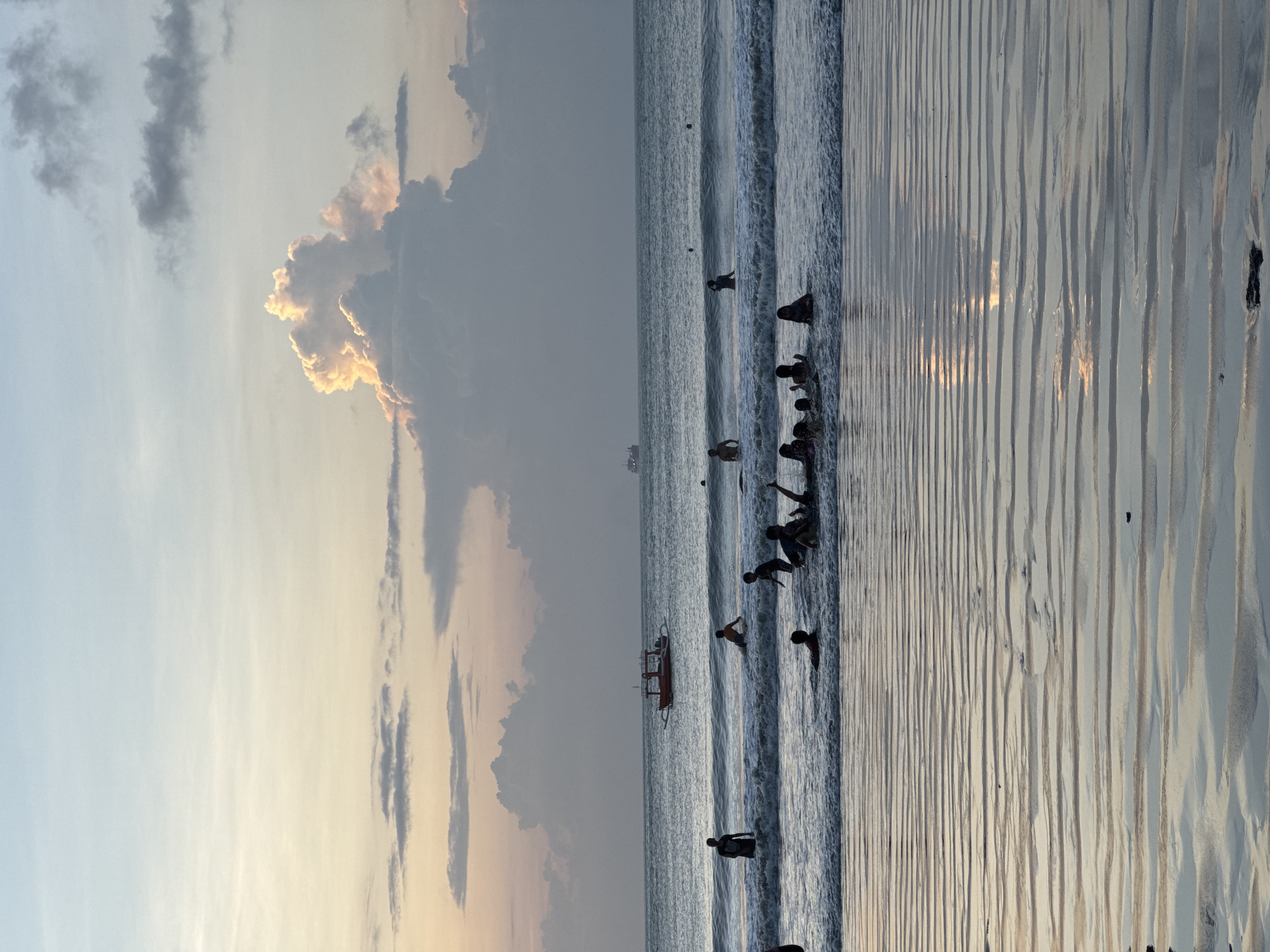 The image shows a beach scene at Kuta Selatan, Indonesia. Several people are in the water near the shore. There is a boat in the water a little further out. The sky is filled with clouds, some of which are illuminated by the setting sun. The sand on the beach is wet and shows patterns from the receding tide.