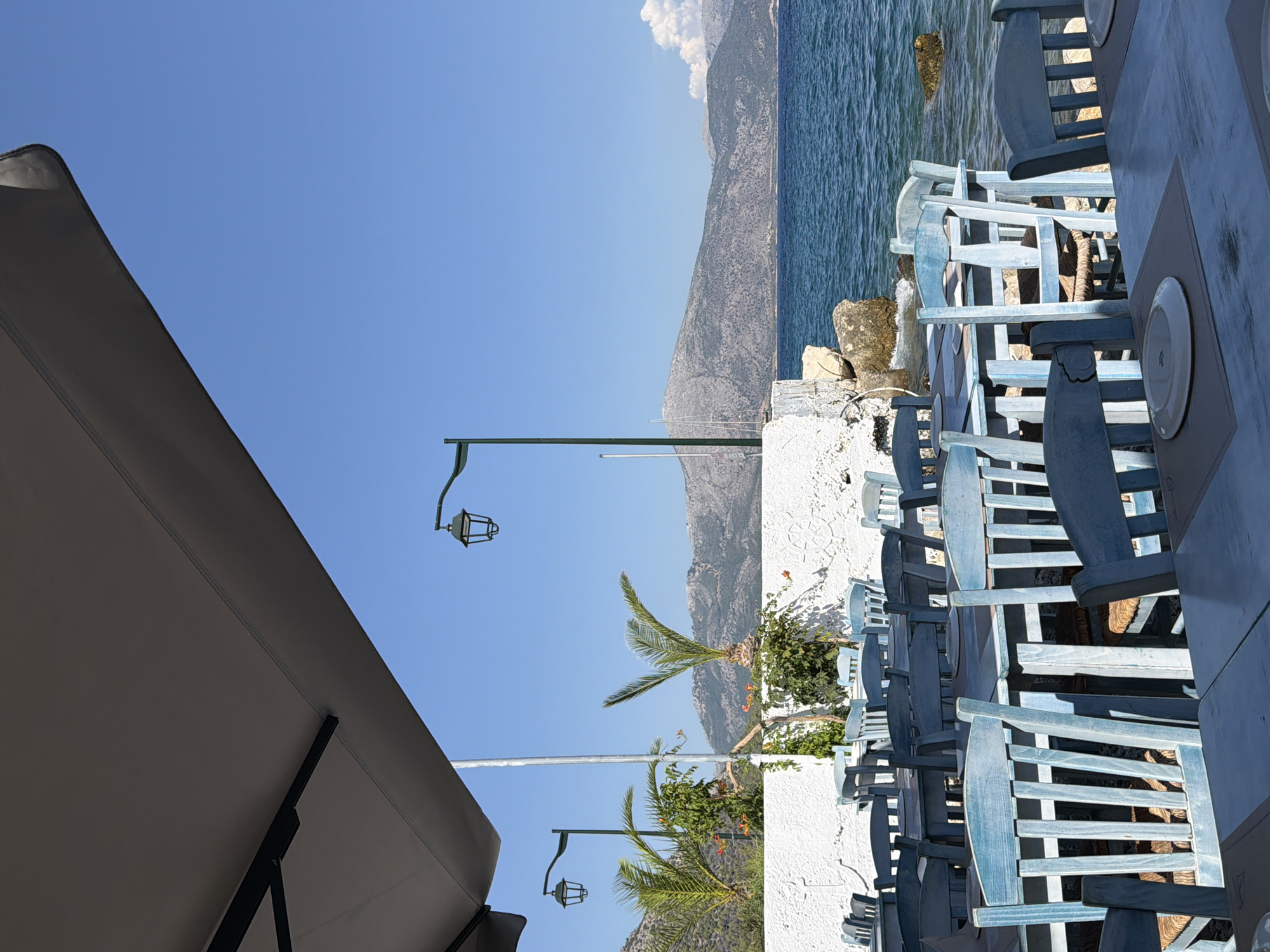 The image depicts an outdoor seating area of a restaurant situated on the waterfront in the Municipal Unit of Kalamos, Greece. In the foreground, several light blue wooden tables and chairs are arranged, some with plates on them. Behind the tables, a low white wall with decorative elements separates the seating area from the sea. Two palm trees and lush green foliage with small orange flowers are visible behind the wall, along with a lamppost. The sea is calm and blue, with a rocky coastline and mountainous terrain in the background. The sky is clear and bright blue. A large dark umbrella shades a portion of the seating area. The overall scene suggests a peaceful, sunny day at a seaside restaurant.