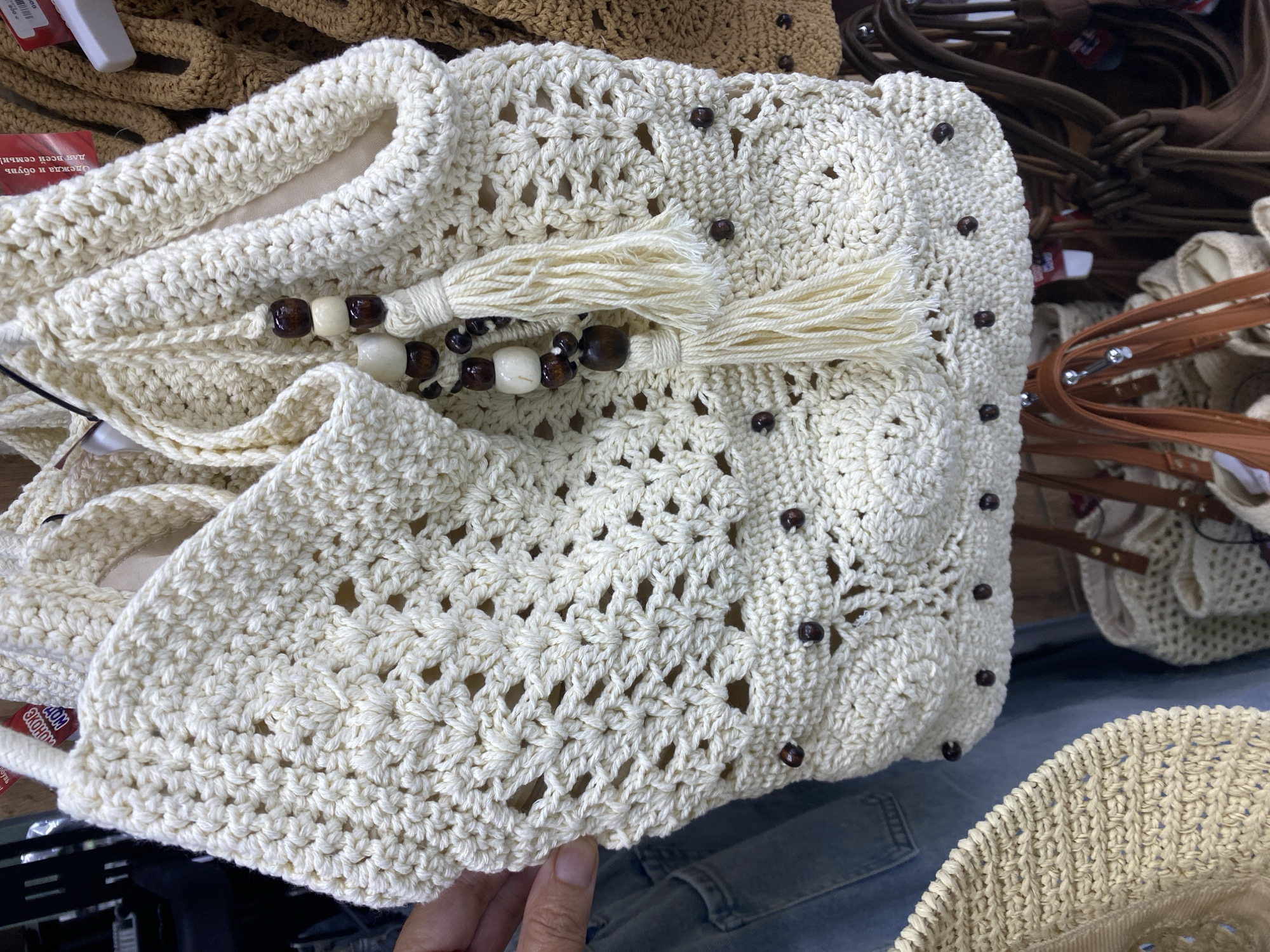 A cream-colored crocheted bag with wooden bead and tassel embellishments is displayed. The bag features an intricate open-weave pattern and is adorned with dark brown beads. It hangs among other bags and clothing items in what appears to be a market stall or boutique. The background shows racks of merchandise and a portion of a person's arm and hand holding the bag. The scene is likely set in Balykchy, Kyrgyzstan, suggesting a commercial or retail environment.