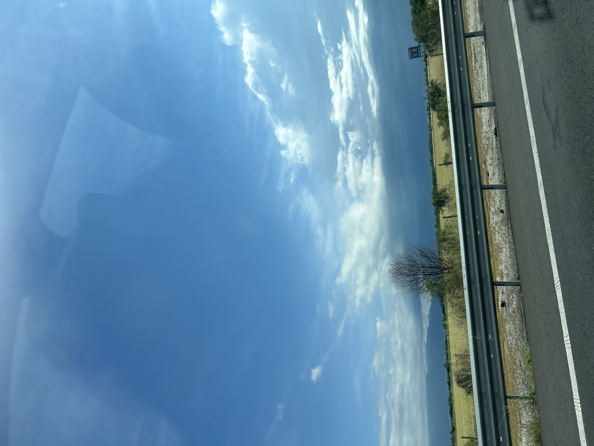 The image displays a sunlit rural landscape observed from a moving vehicle, with the photograph itself oriented vertically (rotated 90 degrees clockwise from its natural landscape perspective). The scene is dominated by a vast blue sky, featuring numerous scattered white cumulus clouds that indicate fair weather, likely during late morning or early afternoon.
On the right side of the image, a paved highway or road surface is visible, marked with a solid white line and a broken white line. A dark metal guardrail runs parallel to the road, separating it from a strip of dry grass and dirt shoulder. Beyond the guardrail, verdant fields stretch into the middle distance, intermingled with some patches of golden vegetation. Several trees are scattered across the landscape, including a prominent bare-branched tree positioned near the guardrail. In the far distance, faint outlines of mountains or hills are visible under a slightly hazy sky.
A blue rectangular sign with white text is discernible in the distance, partly obscured by foliage. The letters "BA" or "BД" appear at the top of the sign, suggesting it is a Bulgarian road sign or advertisement. The top-left corner of the image, when viewed in its current rotation, shows a reflection or glare from a window, consistent with the perspective from inside a vehicle. The overall scene conveys a sense of travel through an open, agricultural area near Malo Konare, Bulgaria.