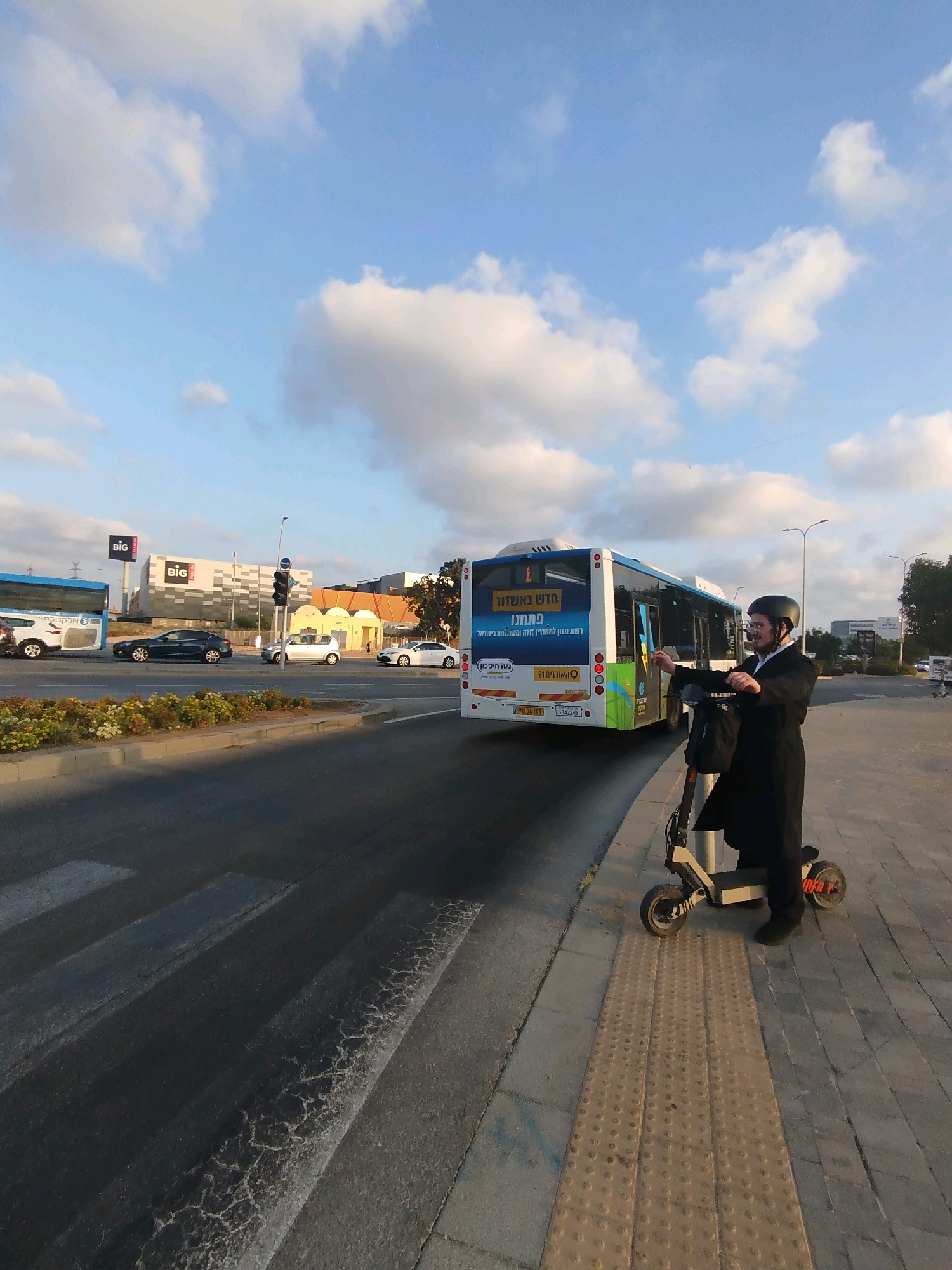 A man wearing traditional Hasidic clothing, including a black coat and hat, is standing on an electric scooter on a sidewalk in Ashdod, Israel. He is wearing a helmet and appears to be interacting with a bus driver or signaling to the bus. The bus, a white and blue public transport vehicle, has Hebrew text on its rear. In the background, there are other vehicles on the road, including cars and another bus, as well as buildings that appear to be commercial establishments. The sky is blue with scattered clouds. The scene takes place at an intersection with a traffic light, which is currently green for traffic moving away from the viewer. The sidewalk has tactile paving.