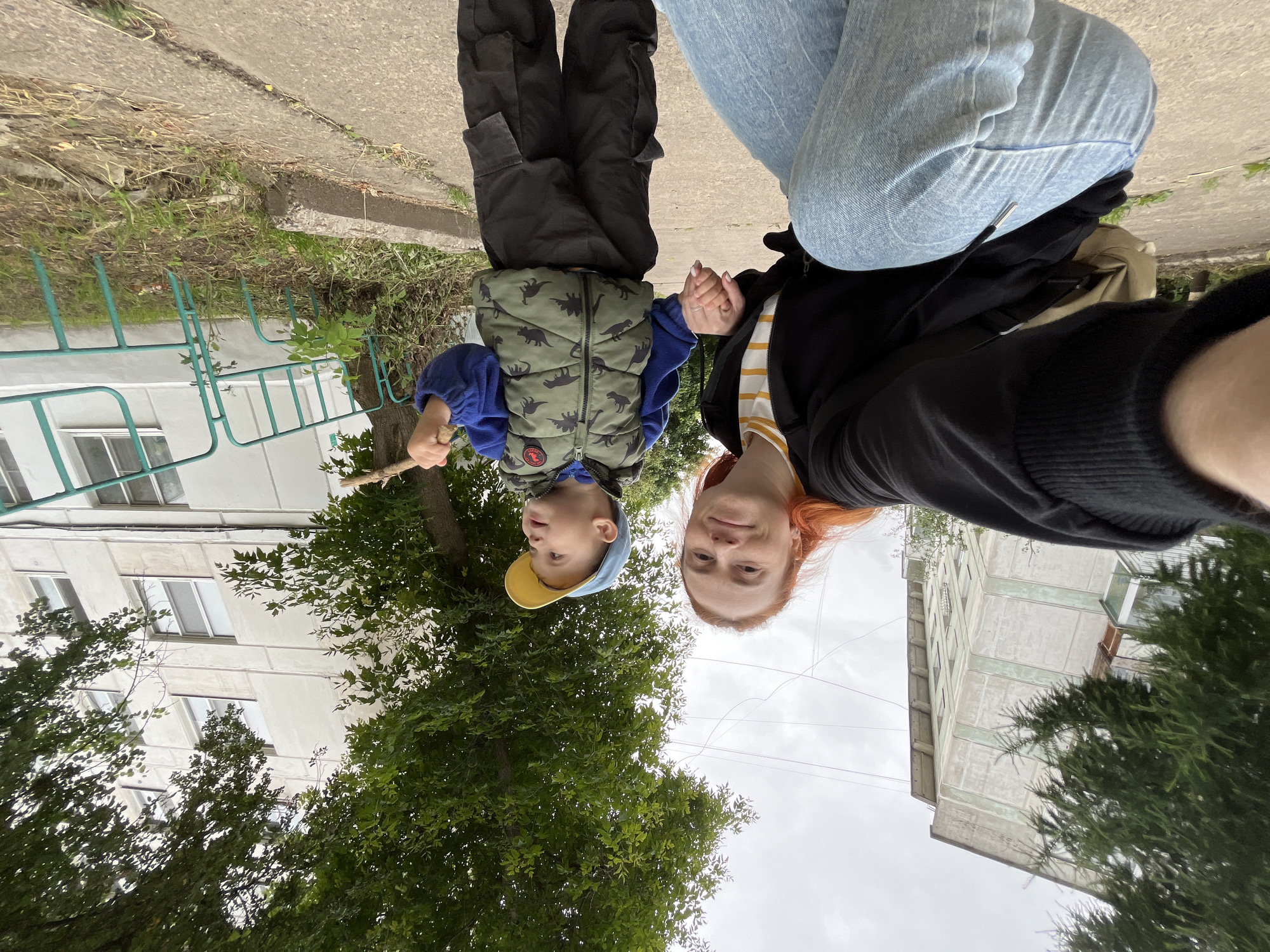 This is a selfie taken outdoors of a woman and a young child, with their heads oriented towards the bottom of the frame. The woman has red hair and is wearing a black jacket over a striped shirt and light blue jeans. She is holding the child's hand. The child is wearing a blue long-sleeved shirt, a camouflage-patterned vest with dinosaur motifs, and a yellow-brimmed hat. He is holding a stick.
The background suggests an urban setting with a building, trees, and a sidewalk. The building appears to be a multi-story apartment building with several windows. There is greenery from trees and grass. The sky is overcast, indicating a cloudy day. The lighting suggests it could be daytime, possibly afternoon, given the diffused light.
The image appears to have been taken in Chaikovsky, Russia. The overall mood seems casual and warm, with the woman and child engaged in an outdoor activity, possibly a walk or playtime. The orientation of the photo suggests it was taken from a low angle, as if the photographer was looking up, or the image has been rotated.