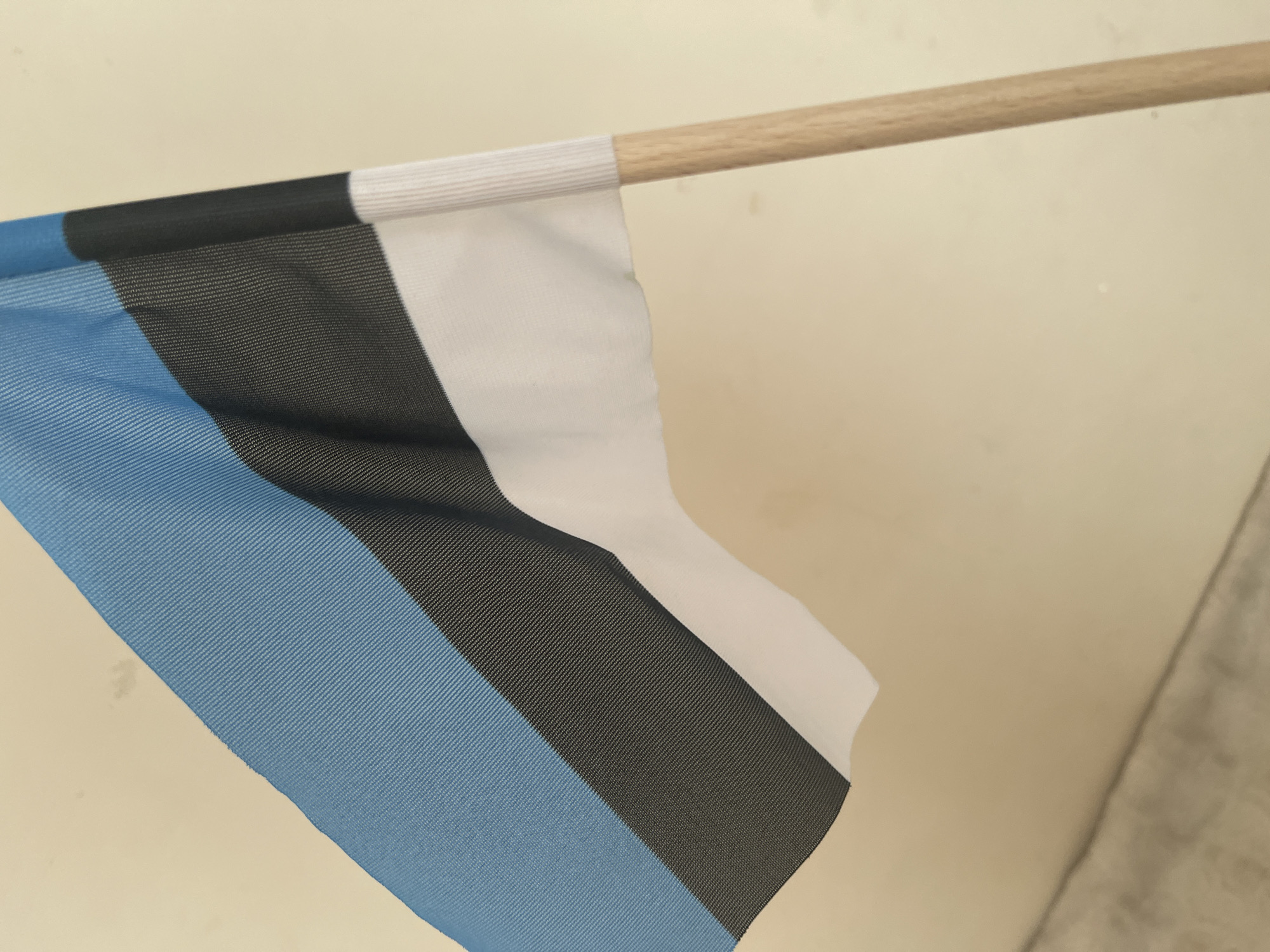The image shows a small flag of Estonia attached to a wooden pole. The flag has three horizontal stripes: blue at the top, black in the middle, and white at the bottom. The flag is partially unfurled and appears to be held against a plain, light-colored wall. The wooden pole is positioned on the right side of the image. The scene is indoors or in a sheltered outdoor space, as suggested by the plain background and lack of natural elements. There is no indication of a specific location in Tallinn, Estonia, in the image.
