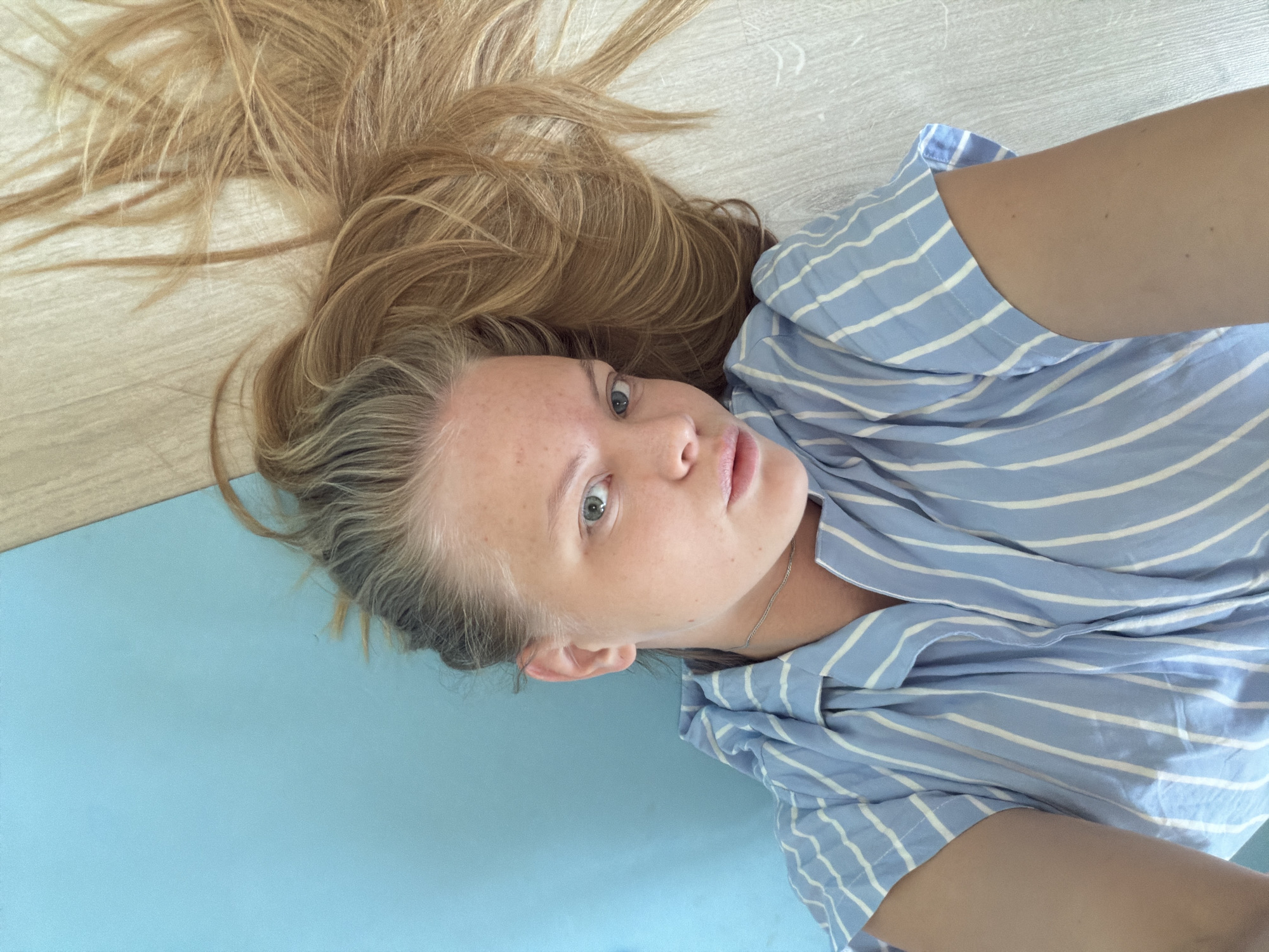 A woman lies on a surface comprised of light blue and light wood-grain sections. She faces upward with light eyes, freckles, and long blonde hair partially spread on the wood-grain section. She wears a blue and white striped garment and a silver necklace. No information connects this image to Izhevsk, Russia.