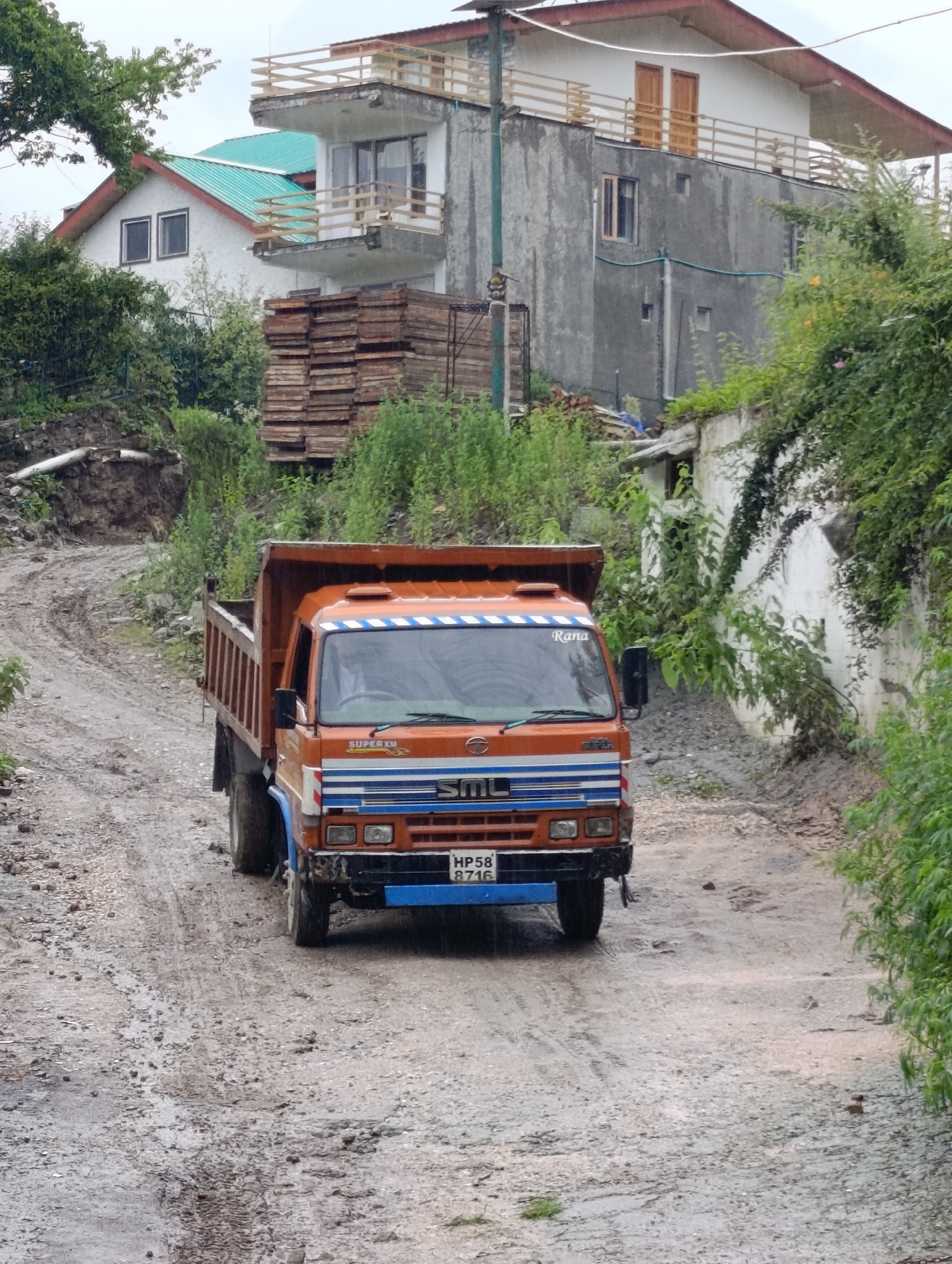 In Aleo, India, an orange dump truck is driving on a wet, muddy dirt road. The truck has a blue bumper and the license plate reads "HP58 B716". The truck is surrounded by lush green vegetation, and in the background, there are two houses. One house is white with a green roof and the other is a gray multi-story building. There are stacks of wood next to the white house. The sky is overcast.