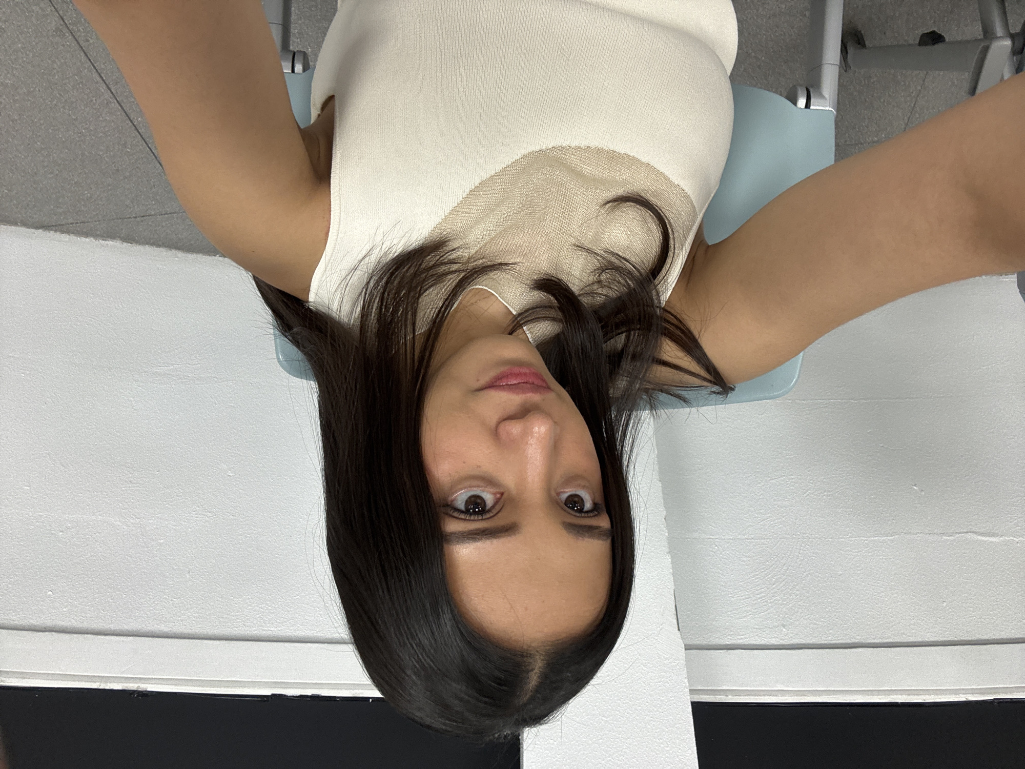 A young female with dark brown, shoulder-length hair and a light tan complexion is depicted in a close-up selfie, looking directly at the camera with a neutral expression. She is wearing a sleeveless, off-white knit top featuring a light brown, semi-sheer mesh panel around the upper chest and collarbone area. Her left arm is partially visible in the lower left foreground, extended towards the camera.
She is situated indoors, specifically within the city of Puebla, Mexico. A light blue chair with a silver-toned metal frame is partially visible behind her on the left. The background consists of a plain, textured white wall with a horizontal molding or ledge running across it. Above this molding, a dark, rectangular area, possibly a window or screen, is visible. The floor is a textured gray material.