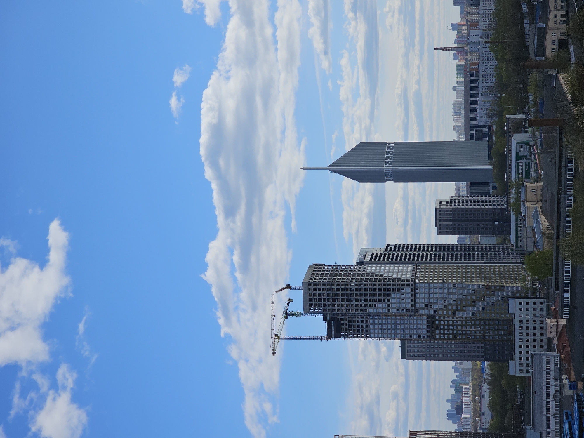 The image displays a sprawling urban landscape in Moscow, Russia, under a clear blue sky dotted with scattered white cumulus clouds. The scene is characterized by a mix of modern high-rise buildings and a denser cityscape in the distance, indicating ongoing development and established urban areas.
In the mid-ground, several multi-story residential or commercial buildings are prominent, with one large structure actively under construction. Two tall tower cranes, one yellow and one green, are positioned beside this building, highlighting the current development phase. To the right of these structures, a striking, dark-colored skyscraper with a distinctive pointed spire reaches high into the sky, serving as a notable architectural landmark.
The foreground and background reveal a rich tapestry of urban infrastructure, including various buildings of different heights and styles, interspersed with green trees. The overall impression is that of a bustling, modern city experiencing significant growth. The time of day appears to be late morning or early afternoon, with bright, natural lighting and fair weather conditions. No discernible text or specific human figures are visible.