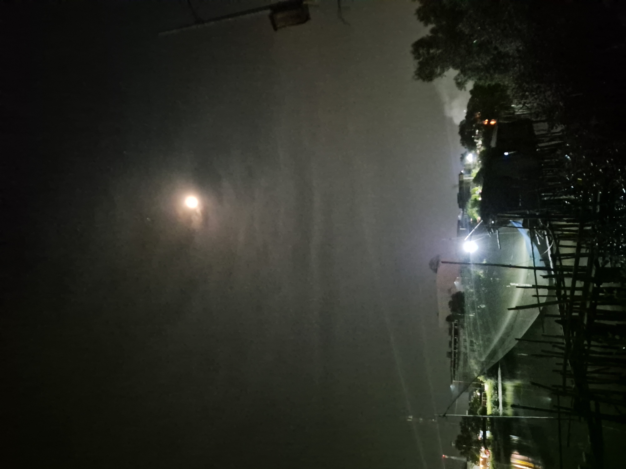 This photograph, taken at night, presents a landscape in Lhokseumawe, Indonesia. The sky is predominantly dark with a visible moon. A body of water reflecting artificial lights and the night sky is present. A structure, possibly a pier or a building on stilts, with a wooden framework extends into the water. Vegetation is visible along the shoreline. Additional lights are scattered in the distance, potentially indicating a residential or urban area. Overall, the scene conveys a sense of quiet activity or settlement on or near the water in a coastal region.