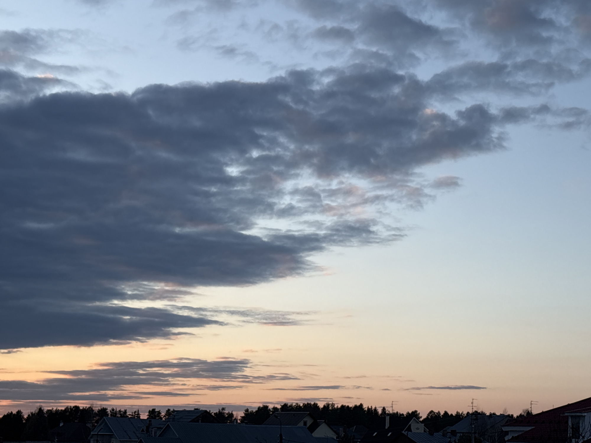 The image depicts a twilight sky over Прикамский, Russia. Dark, heavy clouds dominate the upper left portion of the frame, contrasting with the lighter, pastel-colored sky on the right. Lower in the image, a layer of thin clouds is illuminated by the setting sun. The silhouettes of trees and rooftops of houses are visible along the horizon. Television antennas protrude from the roofs. The overall impression is a calm, quiet evening in a suburban area.